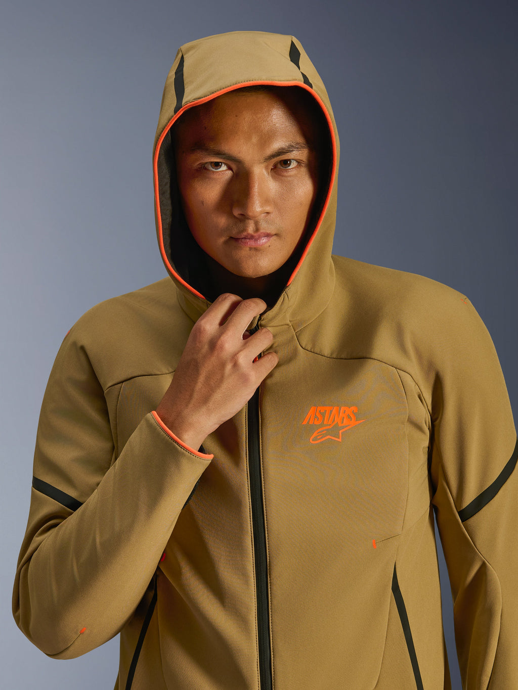 Giacca Alpinestars Aeroshell Waterproof, Dark Khaki, nero e arancione fluo, modello che indossa la giacca con il cappuccio alzato, caratterizzata da vivaci bordi arancione fluo e logo ASTARS arancione, eleganti dettagli neri, softshell impermeabile dal design sportivo moderno