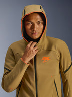 Giacca Alpinestars Aeroshell Waterproof, Dark Khaki, nero e arancione fluo, modello che indossa la giacca con il cappuccio alzato, caratterizzata da vivaci bordi arancione fluo e logo ASTARS arancione, eleganti dettagli neri, softshell impermeabile dal design sportivo moderno