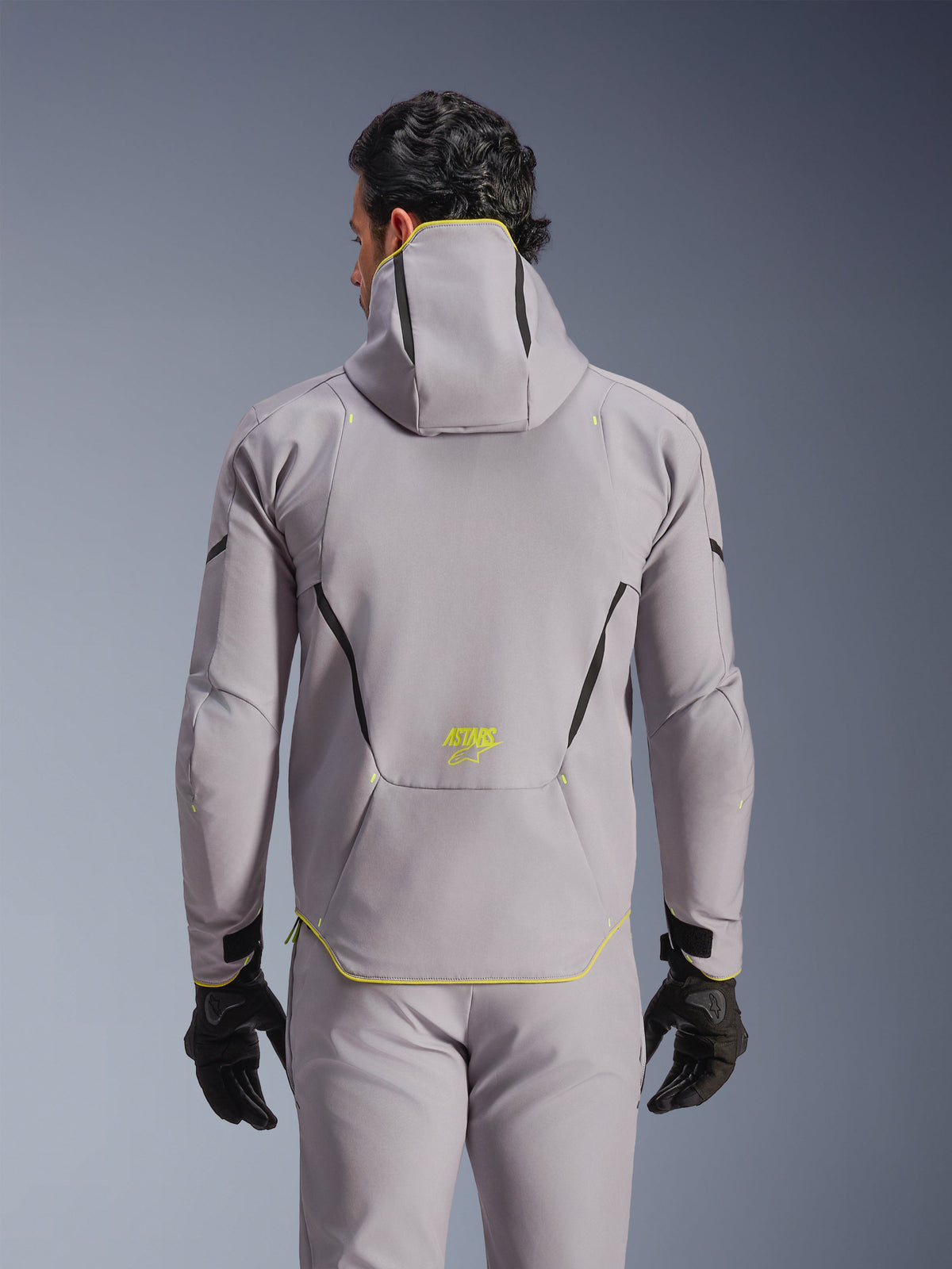 Giacca Alpinestars Aeroshell WP, grigio medio con accenti giallo fluo, vista posteriore con cappuccio integrato e linee di contrasto nere, logo ASTARS giallo sulla parte bassa della schiena, indossata con pantaloni grigi coordinati e guanti neri, giacca da moto softshell sportiva