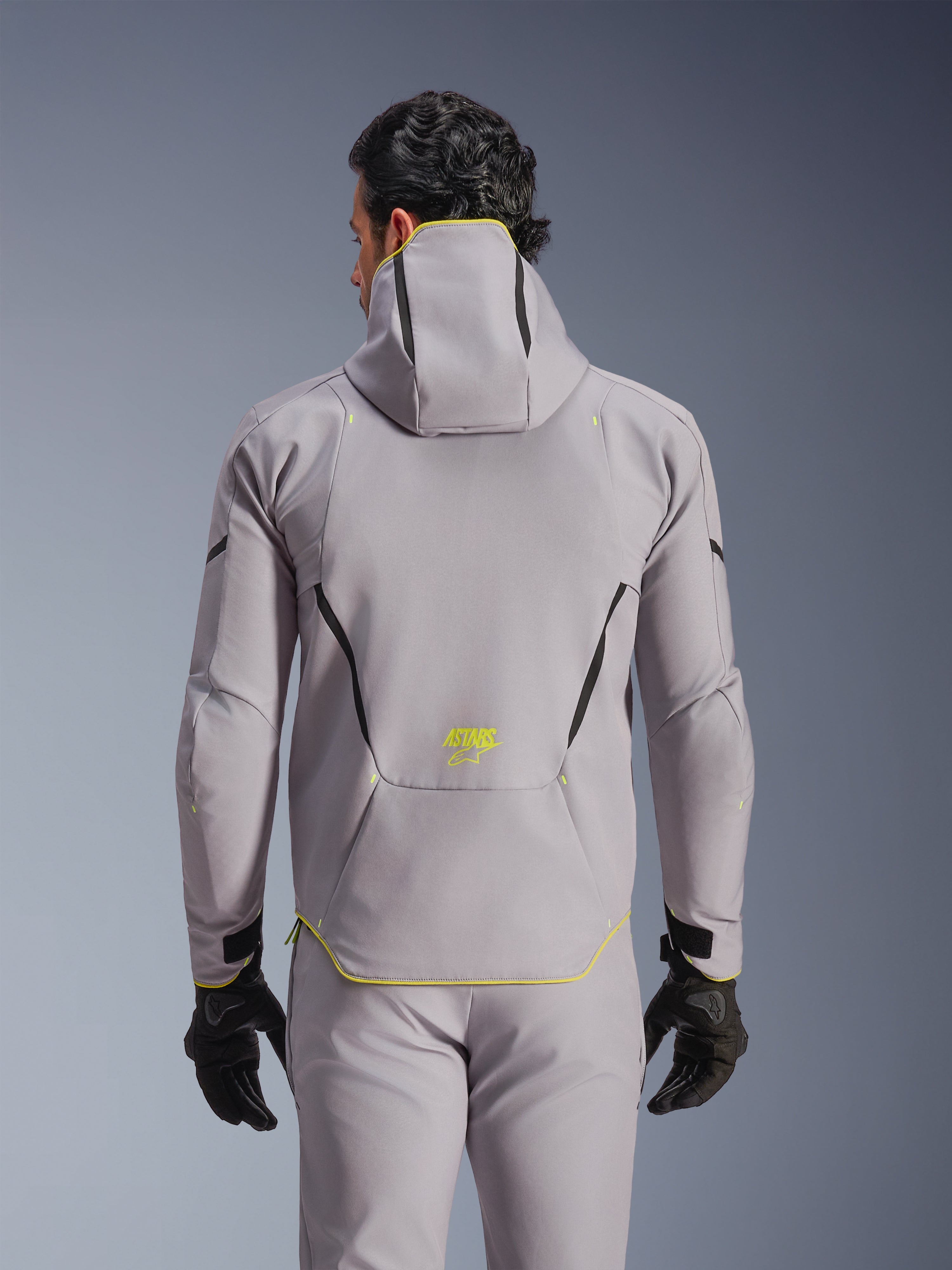 Giacca Alpinestars Aeroshell WP, grigio medio con accenti giallo fluo, vista posteriore con cappuccio integrato e linee di contrasto nere, logo ASTARS giallo sulla parte bassa della schiena, indossata con pantaloni grigi coordinati e guanti neri, giacca da moto softshell sportiva