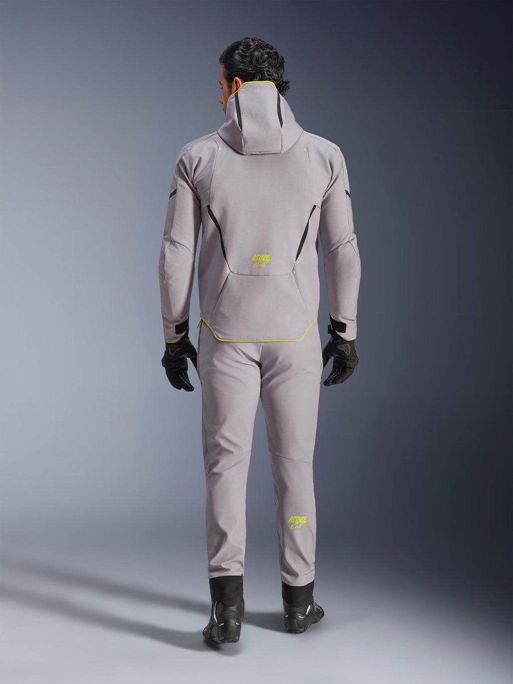 Giacca Alpinestars Aeroshell Waterproof in grigio medio e giallo fluo, vista posteriore completa, indossata con pantaloni grigi coordinati, guanti neri e scarpe da moto nere. Caratterizzata da un design softshell con cappuccio, marchio 'ASTARS' e dettagli giallo fluo, progettata per prestazioni stradali moderne.