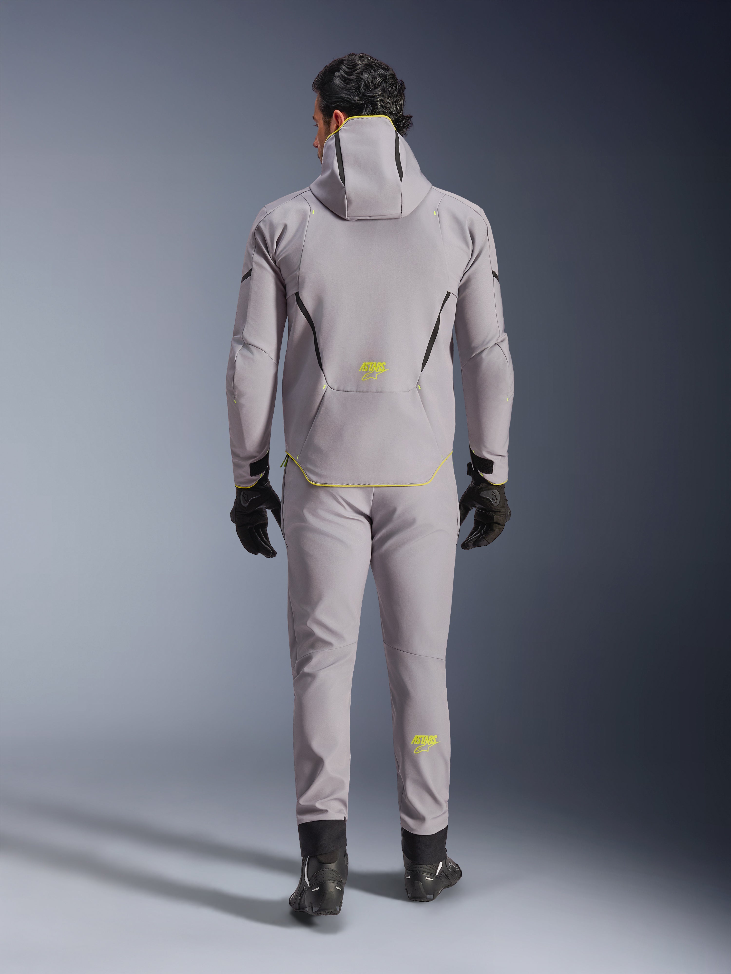 Giacca Alpinestars Aeroshell Waterproof in grigio medio e giallo fluo, vista posteriore completa, indossata con pantaloni grigi coordinati, guanti neri e scarpe da moto nere. Caratterizzata da un design softshell con cappuccio, marchio 'ASTARS' e dettagli giallo fluo, progettata per prestazioni stradali moderne.