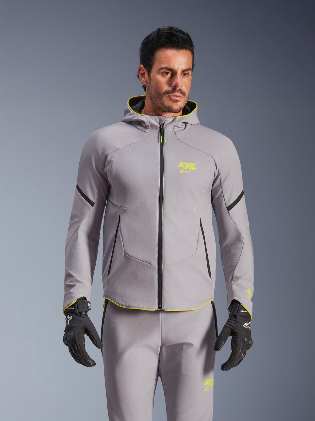 Giacca Alpinestars Aeroshell Waterproof, grigio medio e giallo fluo, giacca da moto softshell con cappuccio e cerniere nere, indossata con pantaloni grigi coordinati e guanti neri, abbigliamento tecnico impermeabile per guida su strada