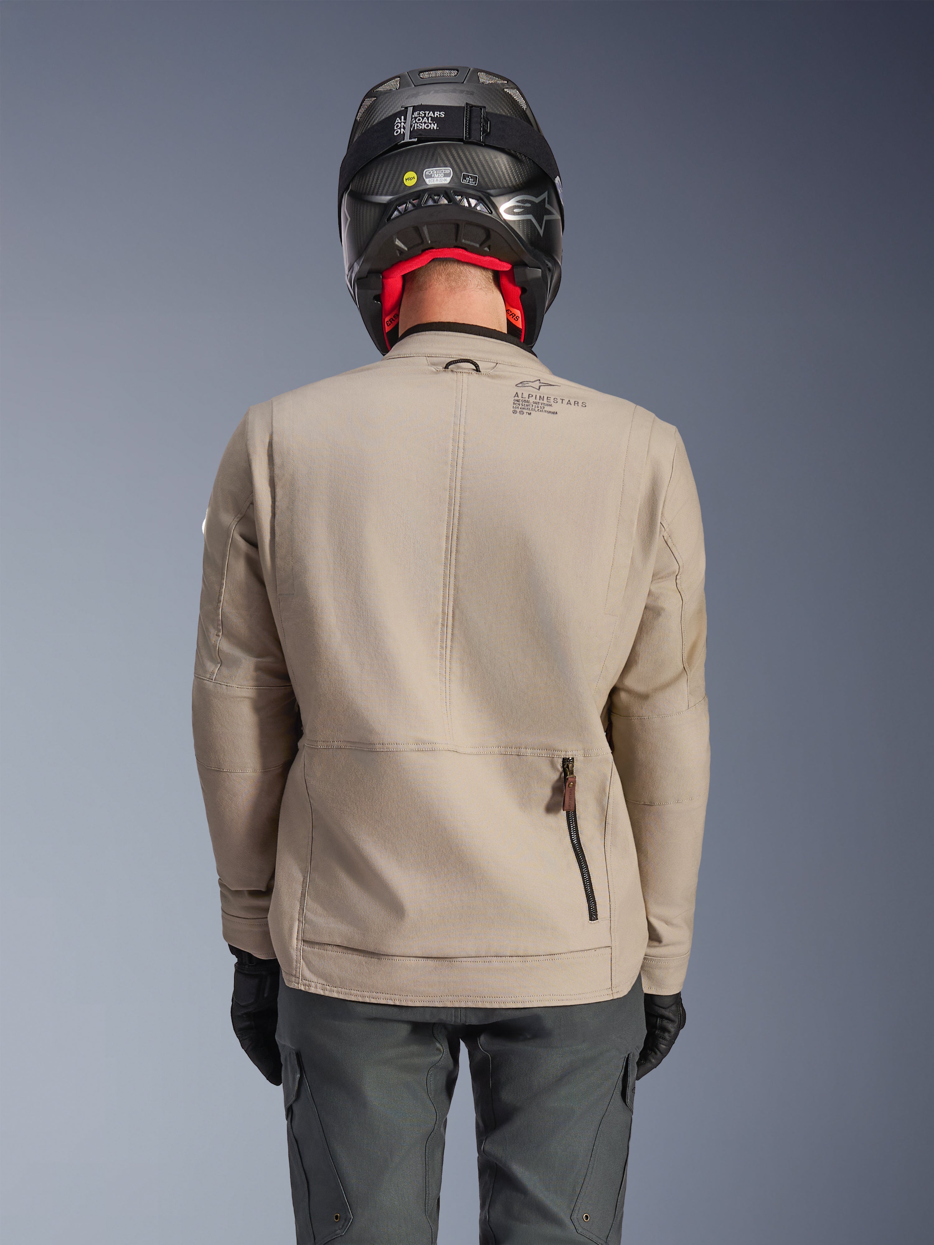 Giacca Alpinestars Borrego Drystar®, giacca da moto adventure, Warm Gray, vista posteriore che mostra la cerniera dello scomparto posteriore e il marchio Alpinestars, indossata con un casco nero e pantaloni da guida grigio antracite