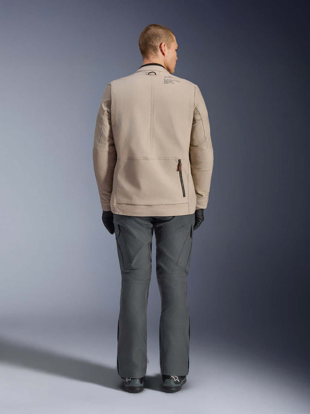 Alpinestars Borrego Drystar® Jacket, vista posteriore di un modello che indossa la giacca da moto in tessuto Warm Gray, abbinata a pantaloni tecnici grigio scuro, guanti neri e stivali, caratterizzata dal design in tela cerata e dai pannelli di espansione posteriori.