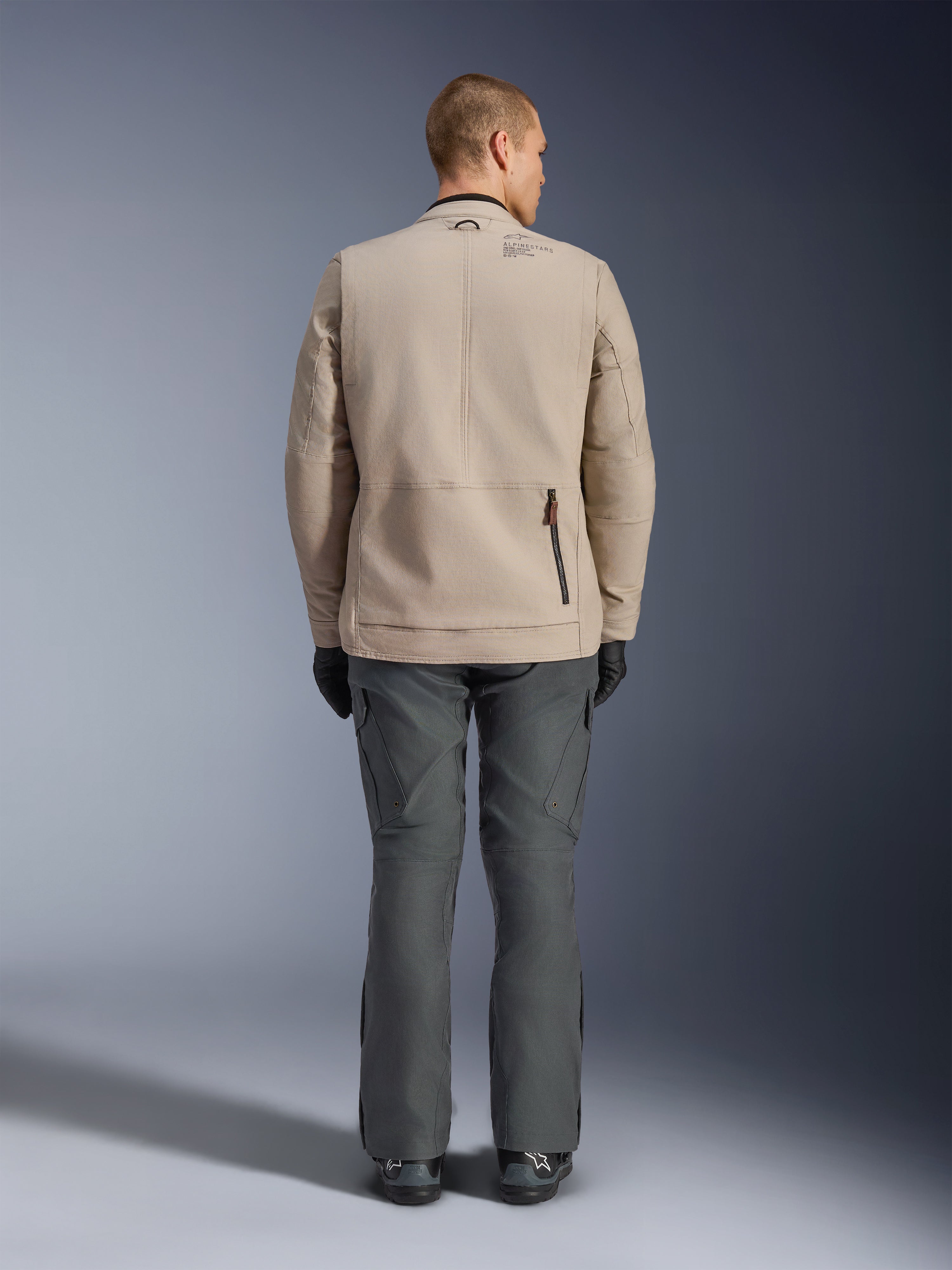 Alpinestars Borrego Drystar® Jacket, vista posteriore di un modello che indossa la giacca da moto in tessuto Warm Gray, abbinata a pantaloni tecnici grigio scuro, guanti neri e stivali, caratterizzata dal design in tela cerata e dai pannelli di espansione posteriori.