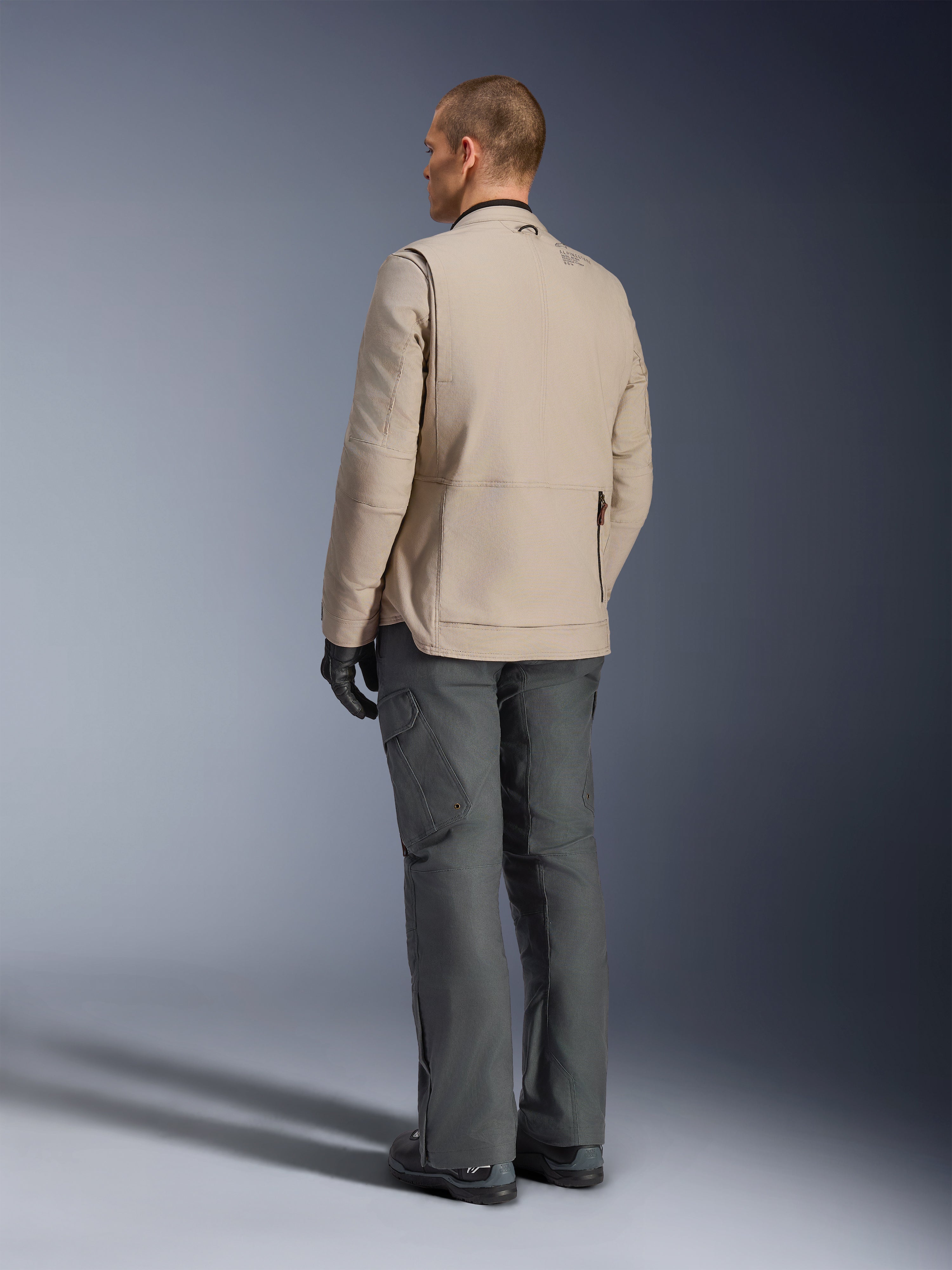 Giacca Alpinestars Borrego Drystar® in Warm Gray, vista posteriore che mostra la robusta texture in tela cerata, i pannelli di espansione sulle spalle e l'ampia tasca posteriore con zip, indossata con pantaloni adventure grigio antracite e guanti neri