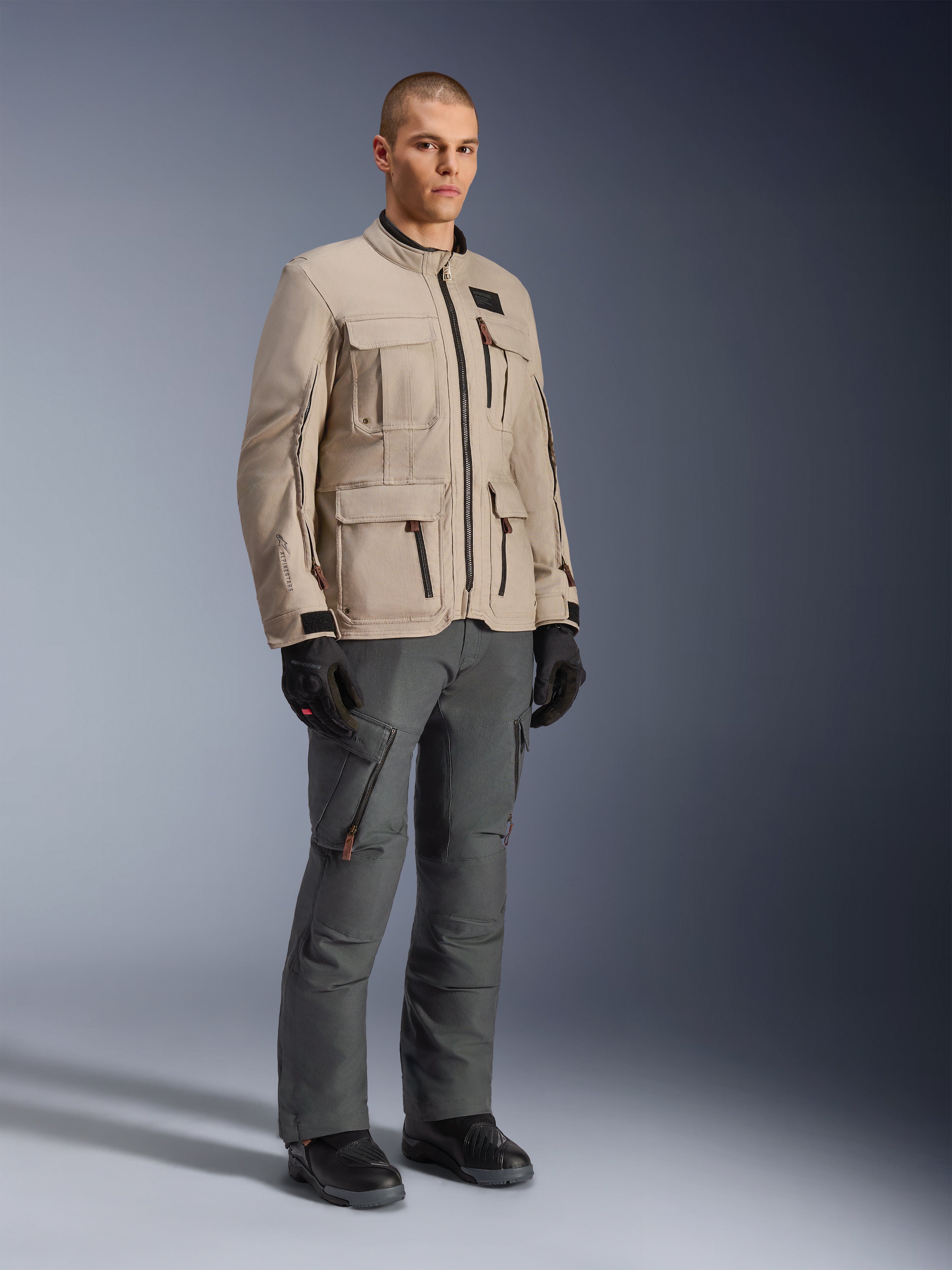 Giacca Alpinestars Borrego Drystar® in Warm Gray, giacca da moto adventure con look in tela cerata e diverse tasche utility, indossata da un modello con pantaloni da moto grigio scuro, guanti neri e stivali neri, ritratto in studio a corpo intero