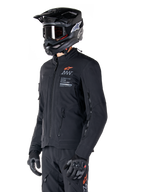 Giacca Alpinestars AMT-8 Stretch Drystar® XF, nera, indossata da un pilota con pantaloni e guanti adventure abbinati, completa di casco da motocross nero e occhiali, aperture di ventilazione con cerniera su petto e braccia, marchio Alpinestars Motion Technology con logo arancione, abbigliamento adventure-touring premium