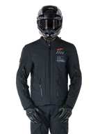 Giacca Alpinestars AMT-8 Stretch Drystar® XF, giacca da moto nera, con logo Alpinestars arancione e marchio AMT grigio, collo alto con cerniera frontale e prese d'aria, indossata con casco e guanti neri, abbigliamento protettivo per adventure touring