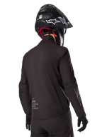 Alpinestars AMT-8 Stretch Drystar® XF Liner, fodera per giacca impermeabile nera, vista posteriore di un pilota che indossa un casco e guanti neri, logo Alpinestars Motion Technology sulla parte bassa della schiena, design minimalista e funzionale per la guida adventure