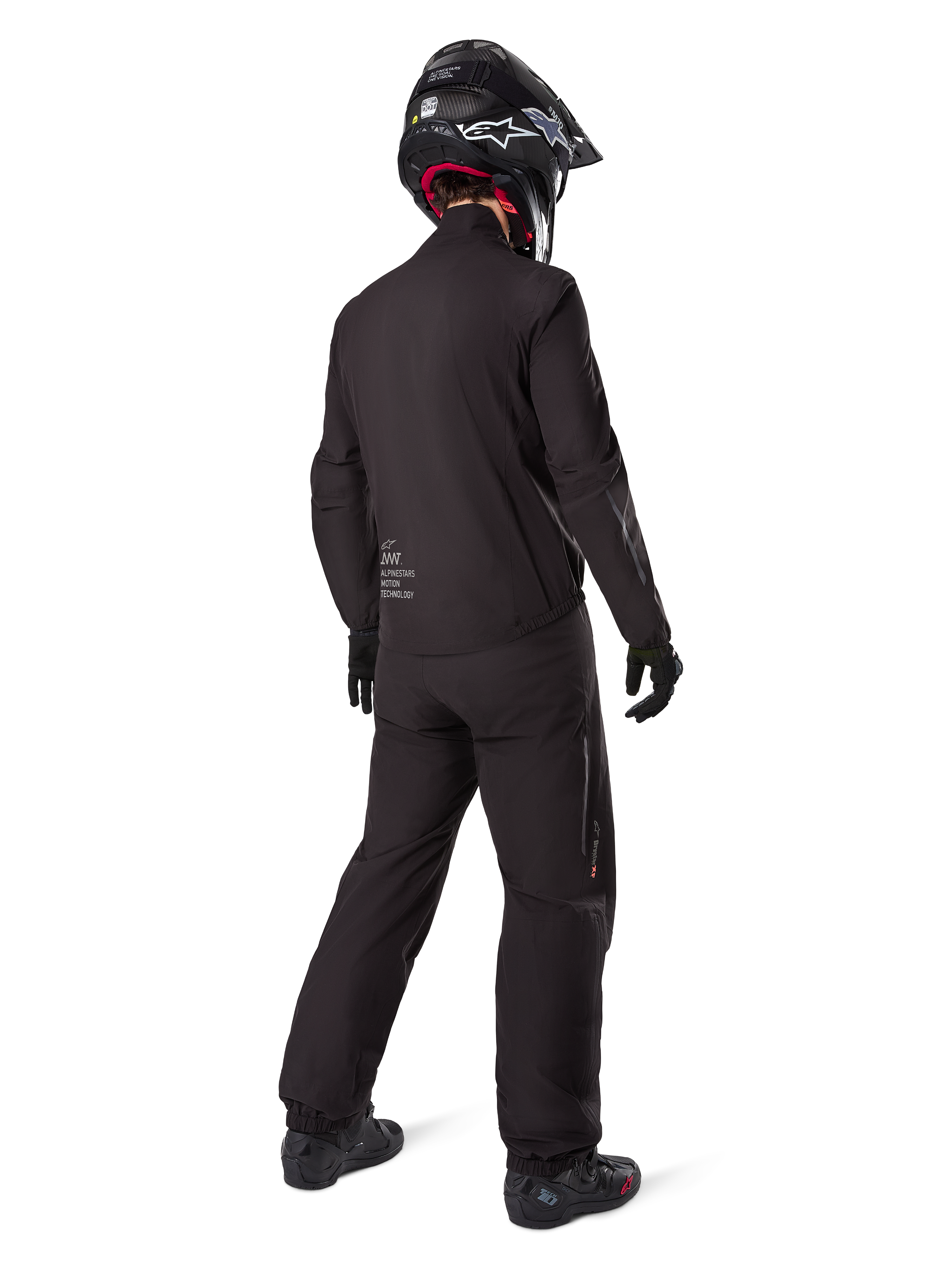 Vista posteriore di un pilota che indossa la fodera della giacca Alpinestars AMT-8 Stretch Drystar® XF, abbigliamento tecnico da moto nero, abbinato a pantaloni coordinati e casco nero, con il marchio "AMT Alpinestars Motion Technology" sul retro, design minimalista per l'adventure touring