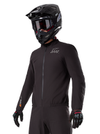 Alpinestars AMT-8 Stretch Drystar® XF Liner, giacca elasticizzata nera antivento e impermeabile, con logo AMT sul petto e marchio Drystar XF sulla manica, indossata con casco adventure nero e guanti protettivi.