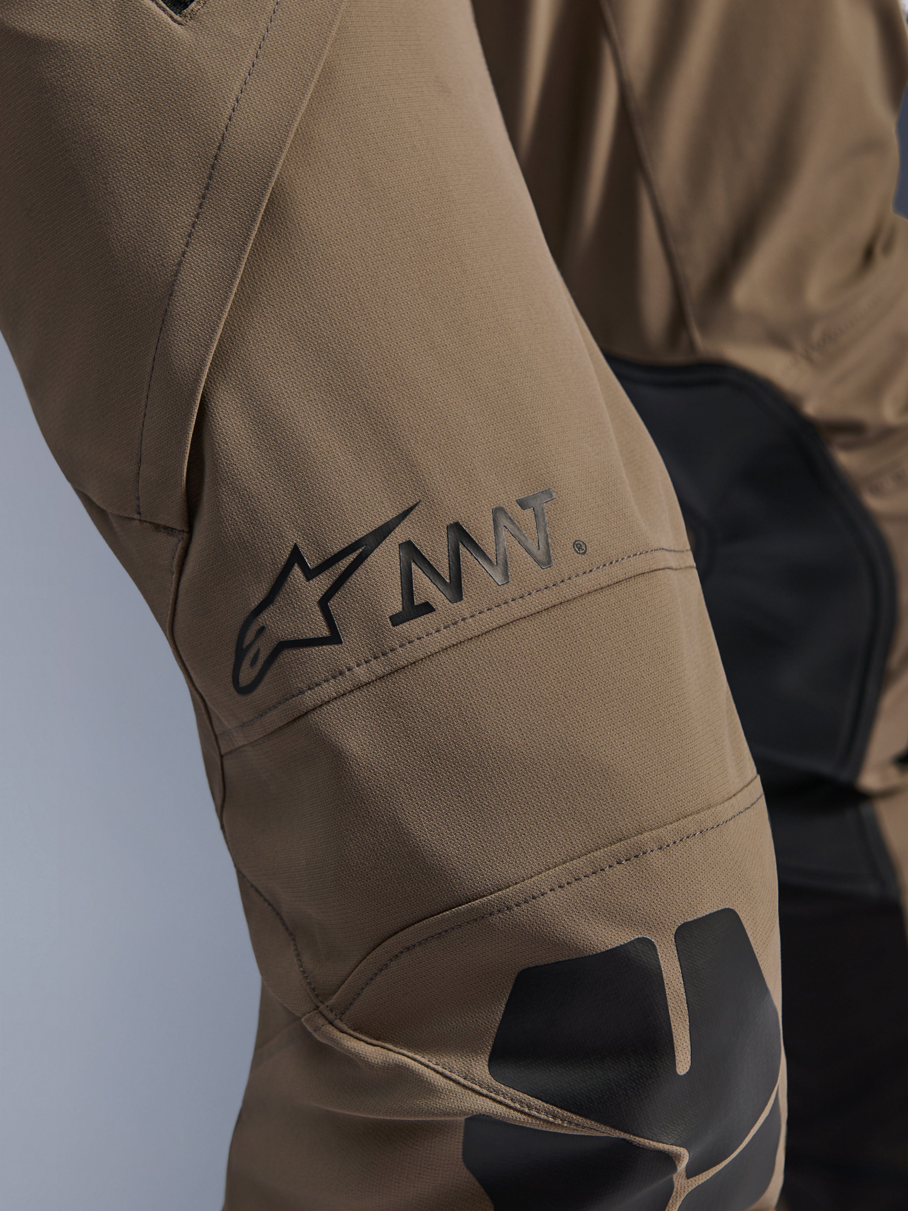 Pantaloni Alpinestars AMT-8 Stretch Drystar® XF in Walnut Black, primo piano della gamba con logo AMT e stella Alpinestars neri, tessuto elasticizzato ad alte prestazioni con inserti neri, abbigliamento tecnico da moto adventure