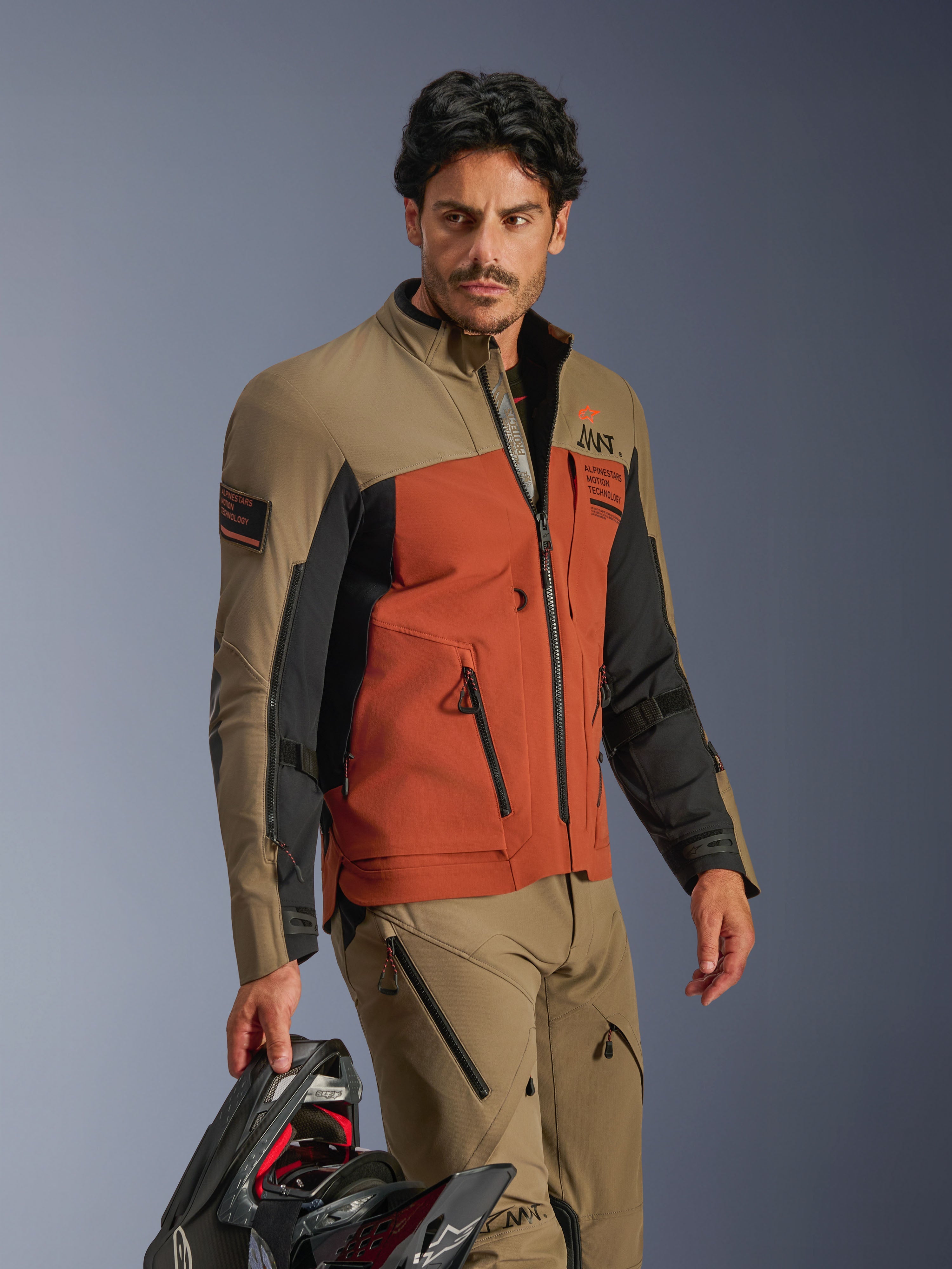 Giacca Alpinestars AMT-8 Stretch Drystar® XF, Walnut Rust Black, giacca da moto adventure in materiale elasticizzato ad alte prestazioni, indossata con pantaloni noce coordinati e con in mano un casco nero, equipaggiamento adventure touring per viaggi a lungo raggio