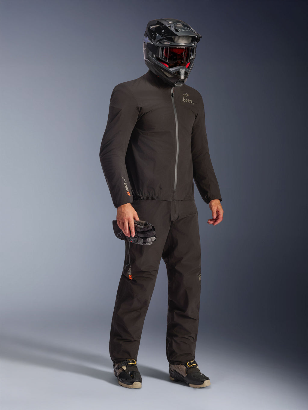 Giacca Alpinestars AMT-8 Stretch Drystar® XF in nero, indossata come parte di un kit da moto completo con pantaloni coordinati e casco nero, caratterizzata da un design in tessuto elegante con logo Drystar XF arancione sulla manica, ideale per l'adventure touring.