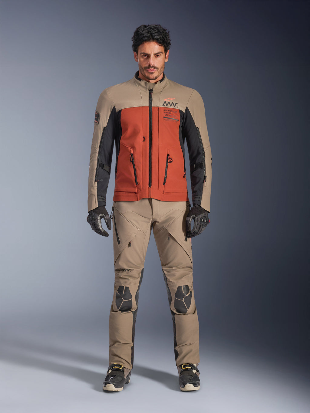 Giacca Alpinestars AMT-8 Stretch Drystar® XF, giacca da moto adventure in colorazione Walnut Rust Black, indossata da un modello con pantaloni tecnici e guanti coordinati, design in tessuto elasticizzato con tasche cargo frontali e loghi, vista frontale.