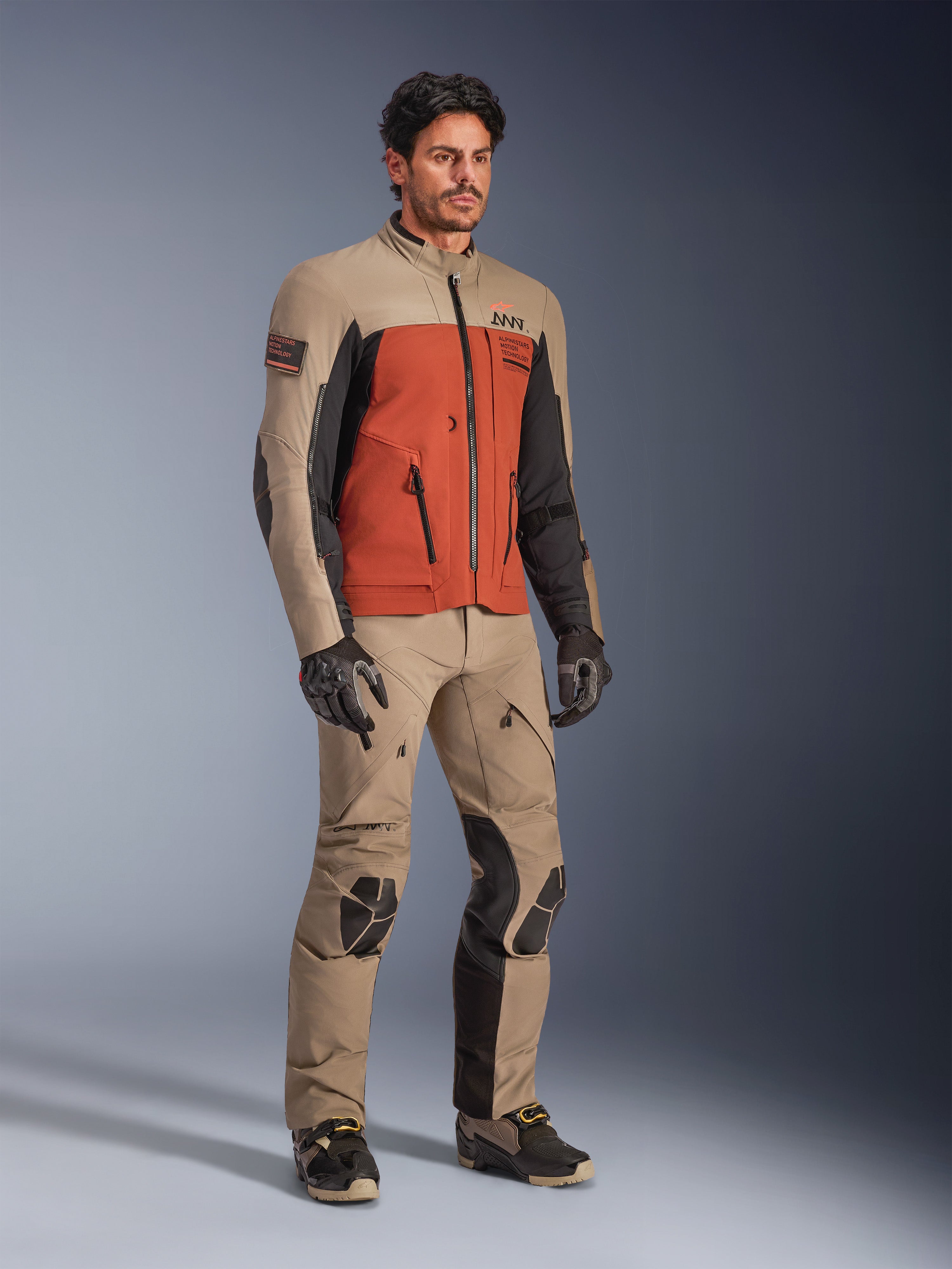 Giacca Alpinestars AMT-8 Stretch Drystar® XF, giacca da moto adventure nella colorazione Walnut Rust Black, indossata con pantaloni e guanti tecnici coordinati, caratterizzata da un design multicolore con pannelli sabbia, arancione ruggine e nero, ideale per il mototurismo a lungo raggio.
