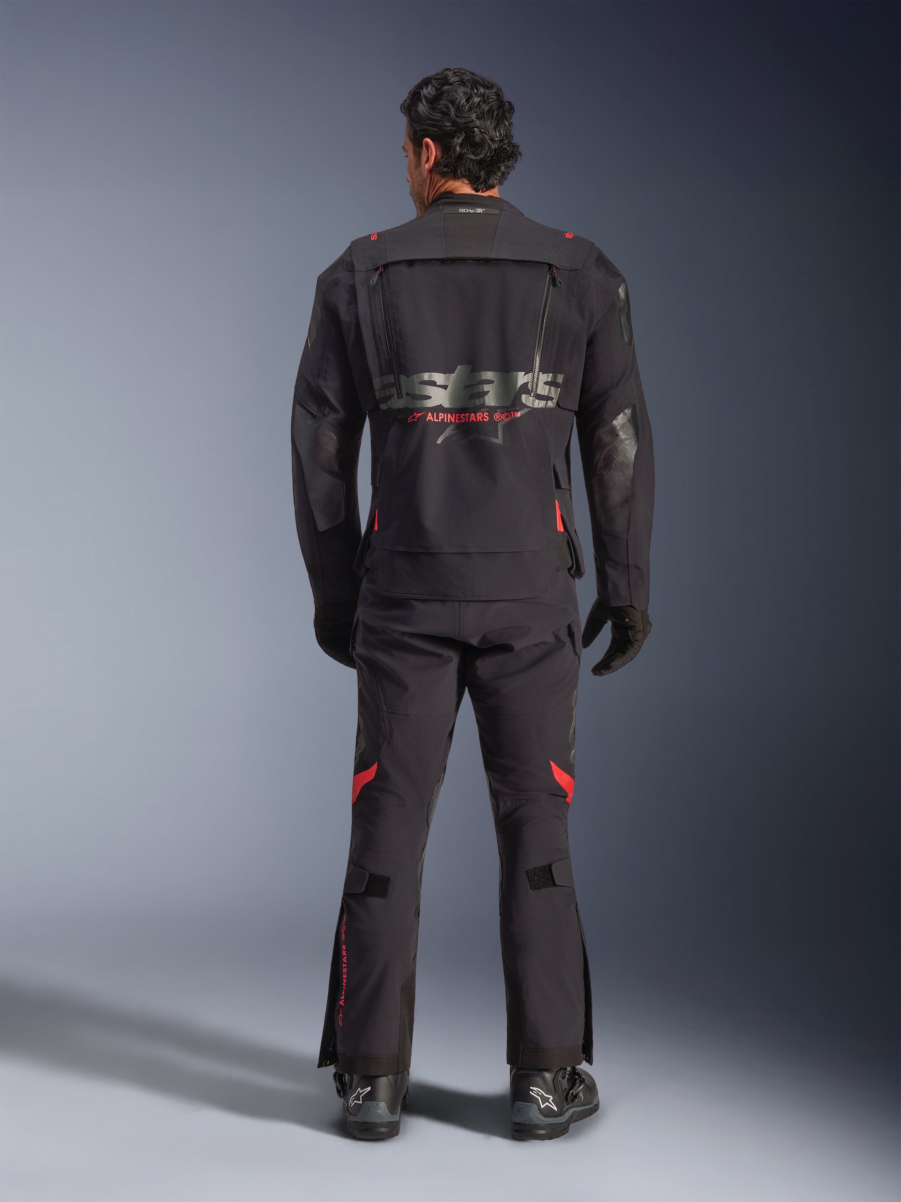 Un pilota visto da dietro che indossa la giacca laminata Alpinestars Halo Pro Drystar® XF in Black Dark Shadow, con un grande logo Astars grigio e marchio Alpinestars rosso, pannelli di ventilazione modulari sulla schiena, abbinata a pantaloni da adventure coordinati e stivali neri.