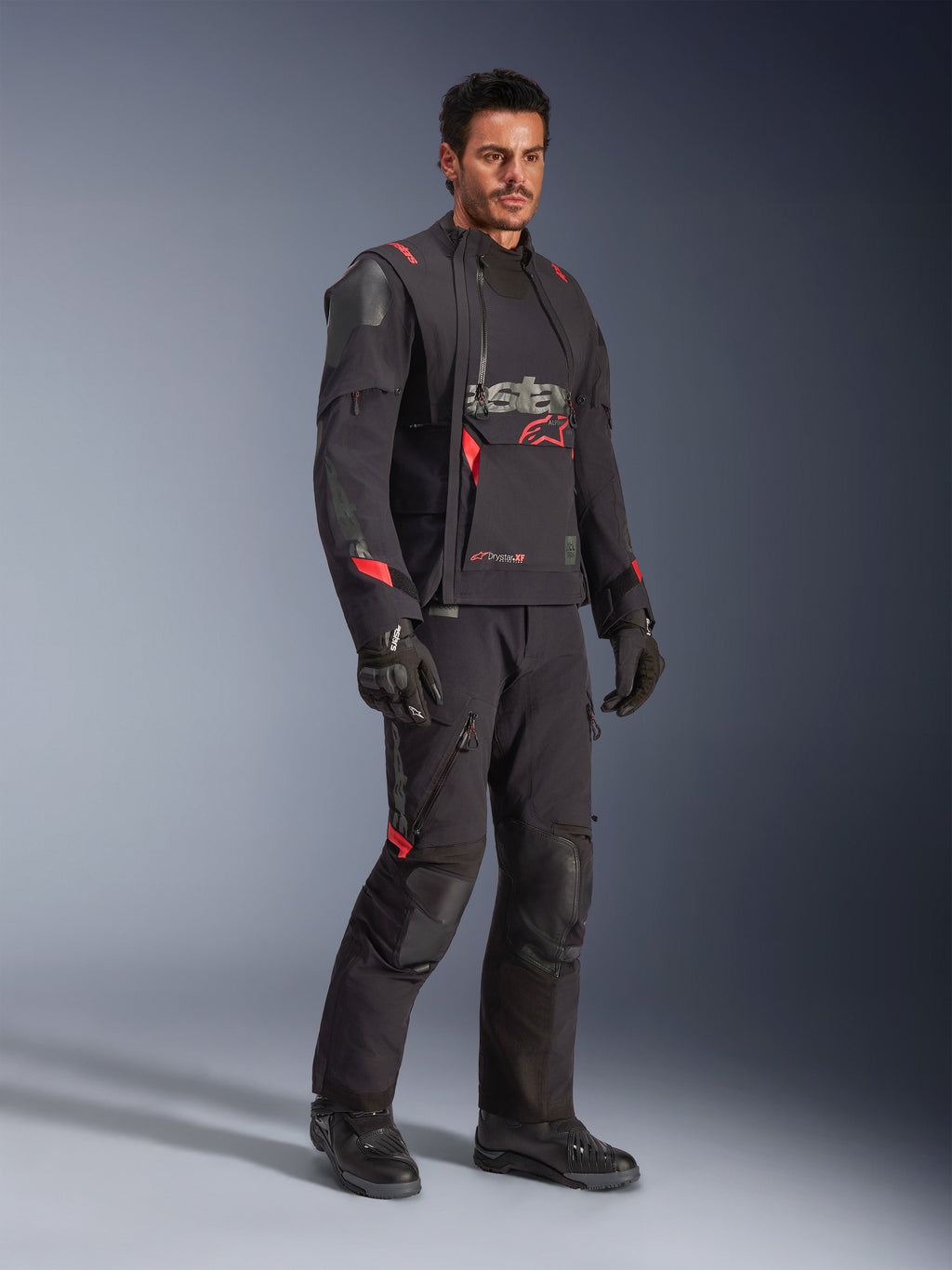 Motociclista con giacca Alpinestars Halo Pro Drystar® XF Laminated in Black Dark Shadow, abbigliamento adventure completo con pantaloni, guanti e stivali coordinati, pannelli frontali asimmetrici modulari e accenti rossi del marchio