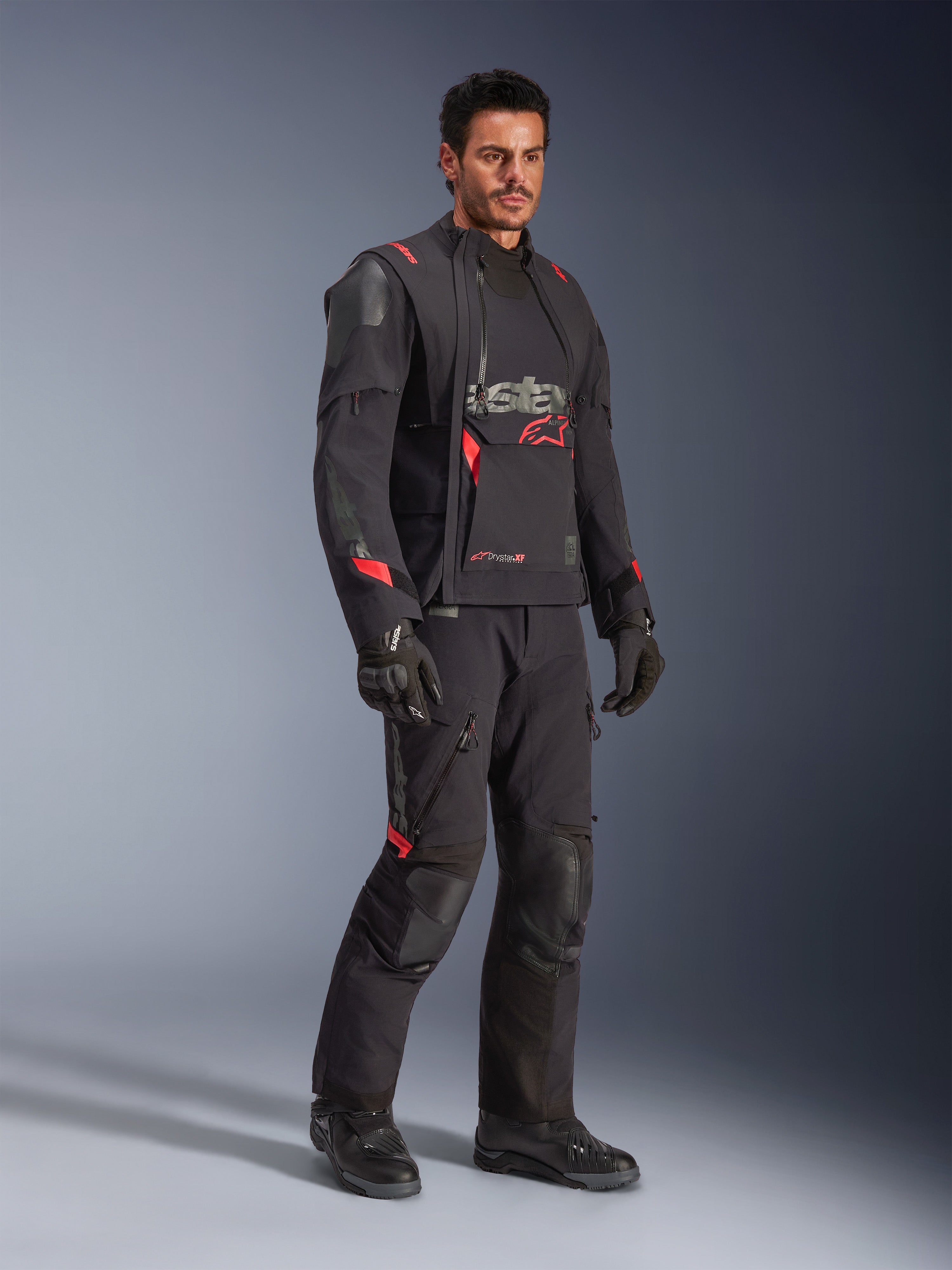Motociclista con giacca Alpinestars Halo Pro Drystar® XF Laminated in Black Dark Shadow, abbigliamento adventure completo con pantaloni, guanti e stivali coordinati, pannelli frontali asimmetrici modulari e accenti rossi del marchio