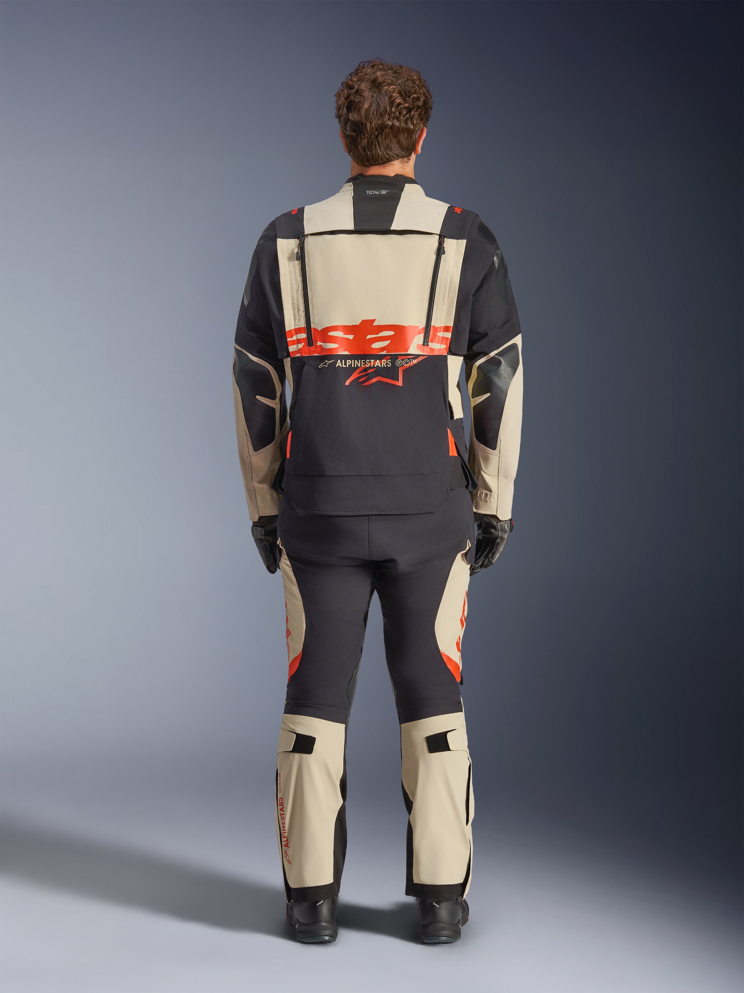 Giacca laminata Alpinestars Halo Pro Drystar® XF, giacca da moto adventure, Mastic Black con logo arancione, vista posteriore che mostra il grande marchio sulla schiena e le cerniere di ventilazione modulari, indossata con pantaloni adventure coordinati per prestazioni su ogni terreno