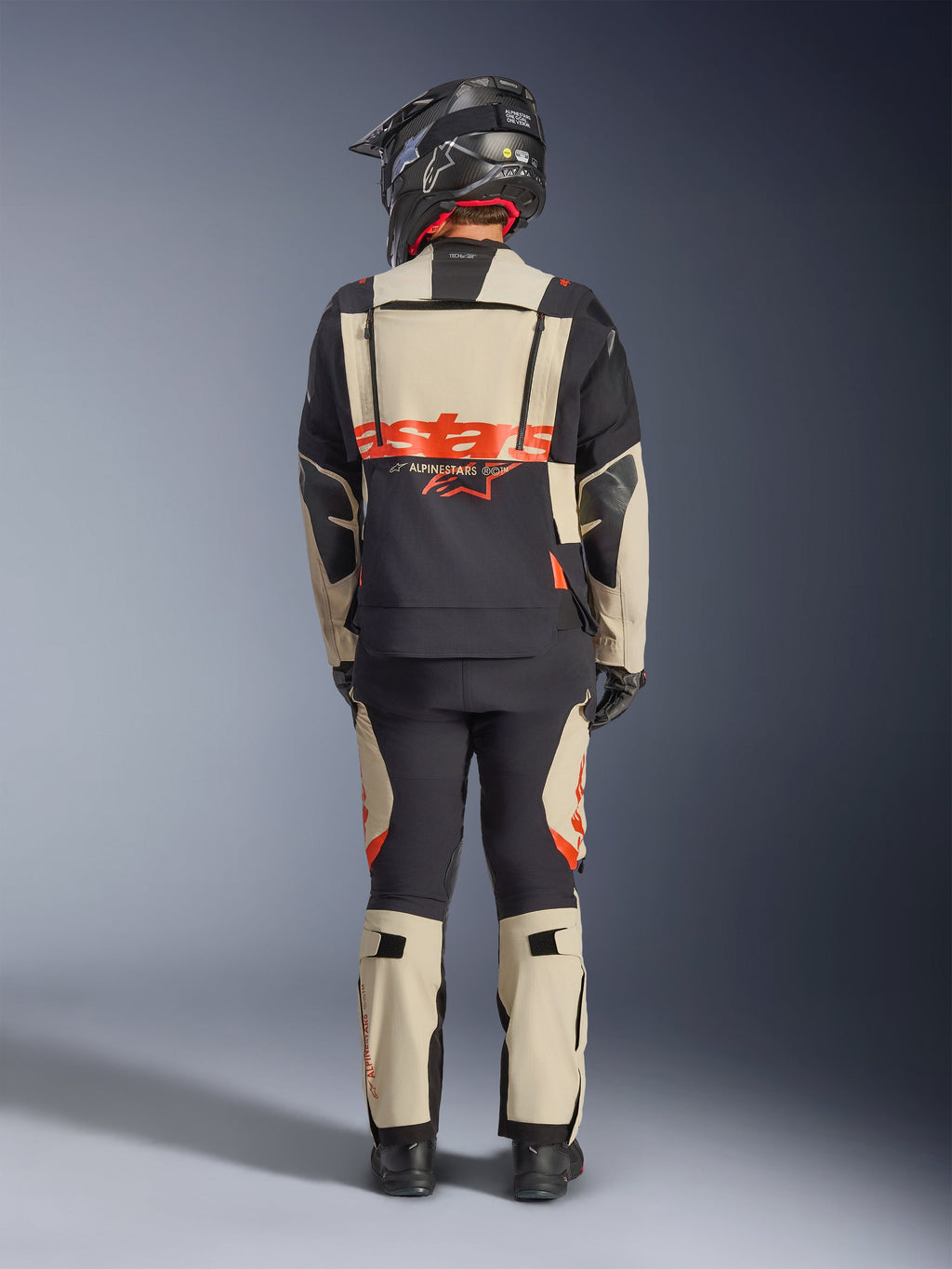 Giacca laminata Alpinestars Halo Pro Drystar® XF, giacca da moto adventure, Mastic Black, vista posteriore di un pilota che indossa pantaloni coordinati e un casco nero, con branding Alpinestars arancione acceso e pannelli di ventilazione modulari per una guida adventure versatile.
