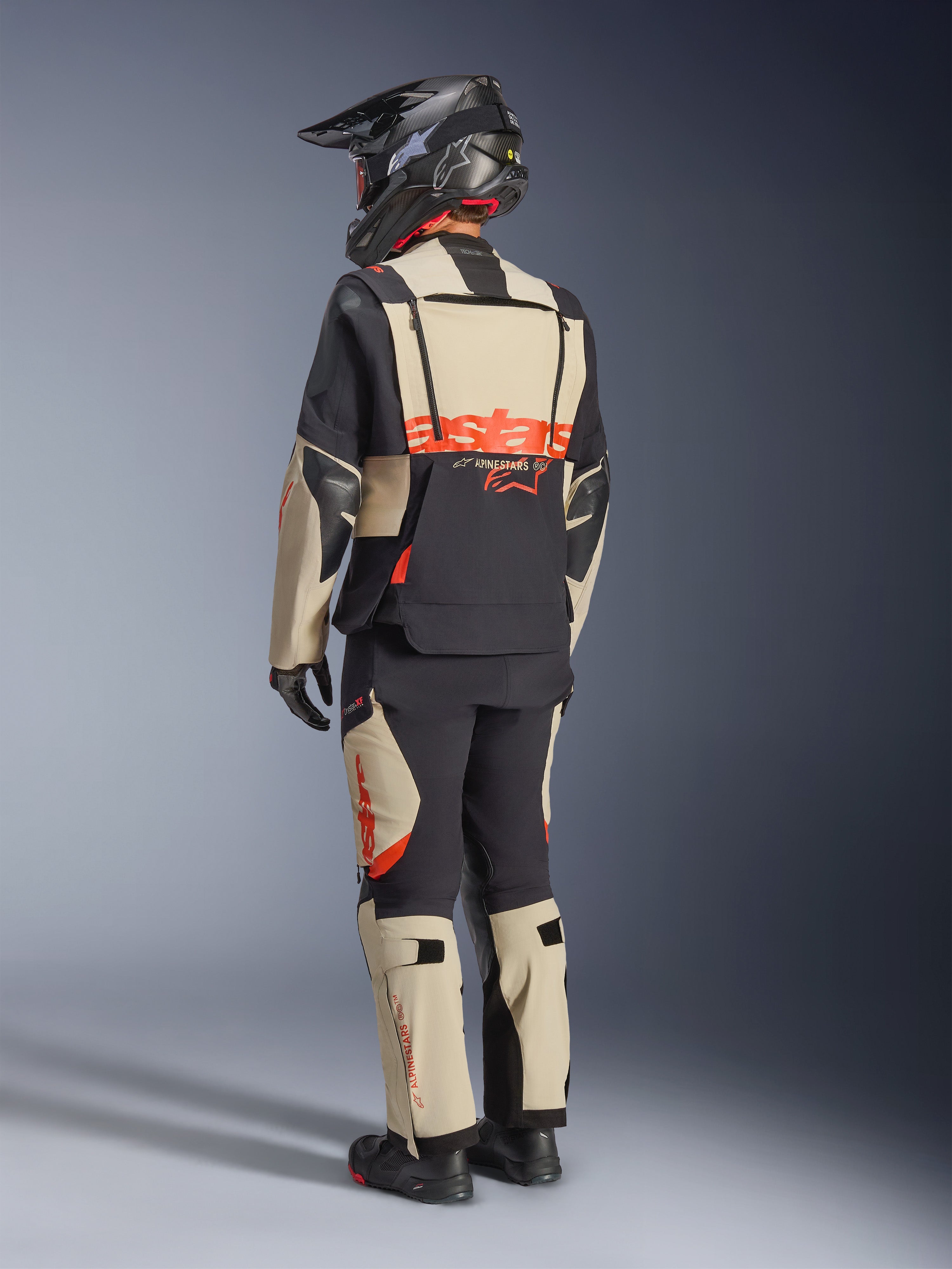Giacca laminata Alpinestars Halo Pro Drystar® XF, Mastic Black, vista posteriore di un pilota che indossa la tuta completa da moto adventure con pantaloni coordinati, casco adventure nero e stivali, con un grande logo Alpinestars rosso sulla schiena.