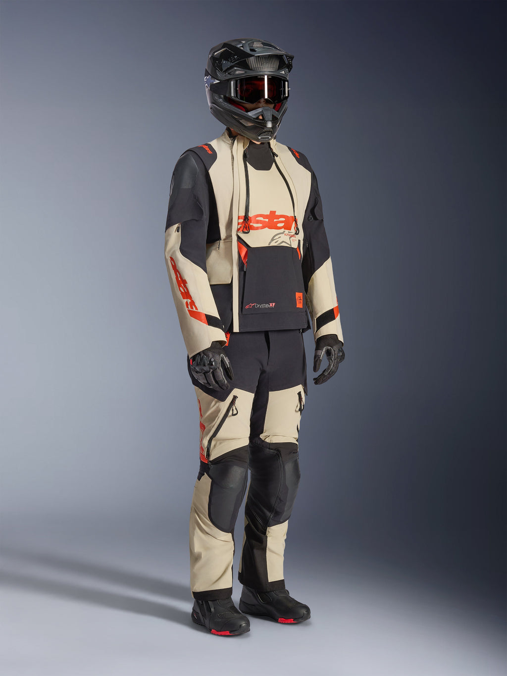 Alpinestars Halo Pro Drystar® XF Laminated Jacket in Mastic Black, giacca da moto adventure indossata con pantaloni coordinati, casco integrale, guanti e stivali, caratterizzata da pannelli beige e neri con loghi rossi, design tecnico multistagione.