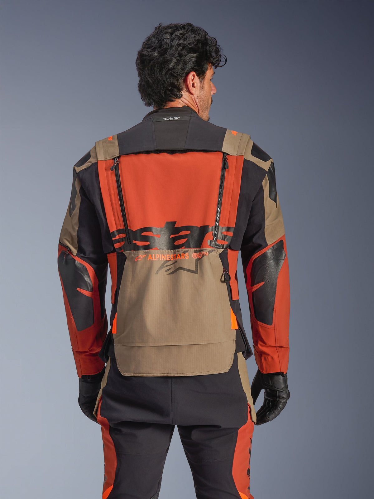 Giacca laminata Alpinestars Halo Pro Drystar® XF, colorazione Walnut Rust Black, vista posteriore, con ampio pannello posteriore arancione, marchio ASTARS nero, tasca portaoggetti inferiore walnut e colletto Tech-Air® Ready, indossata con pantaloni adventure coordinati e guanti neri