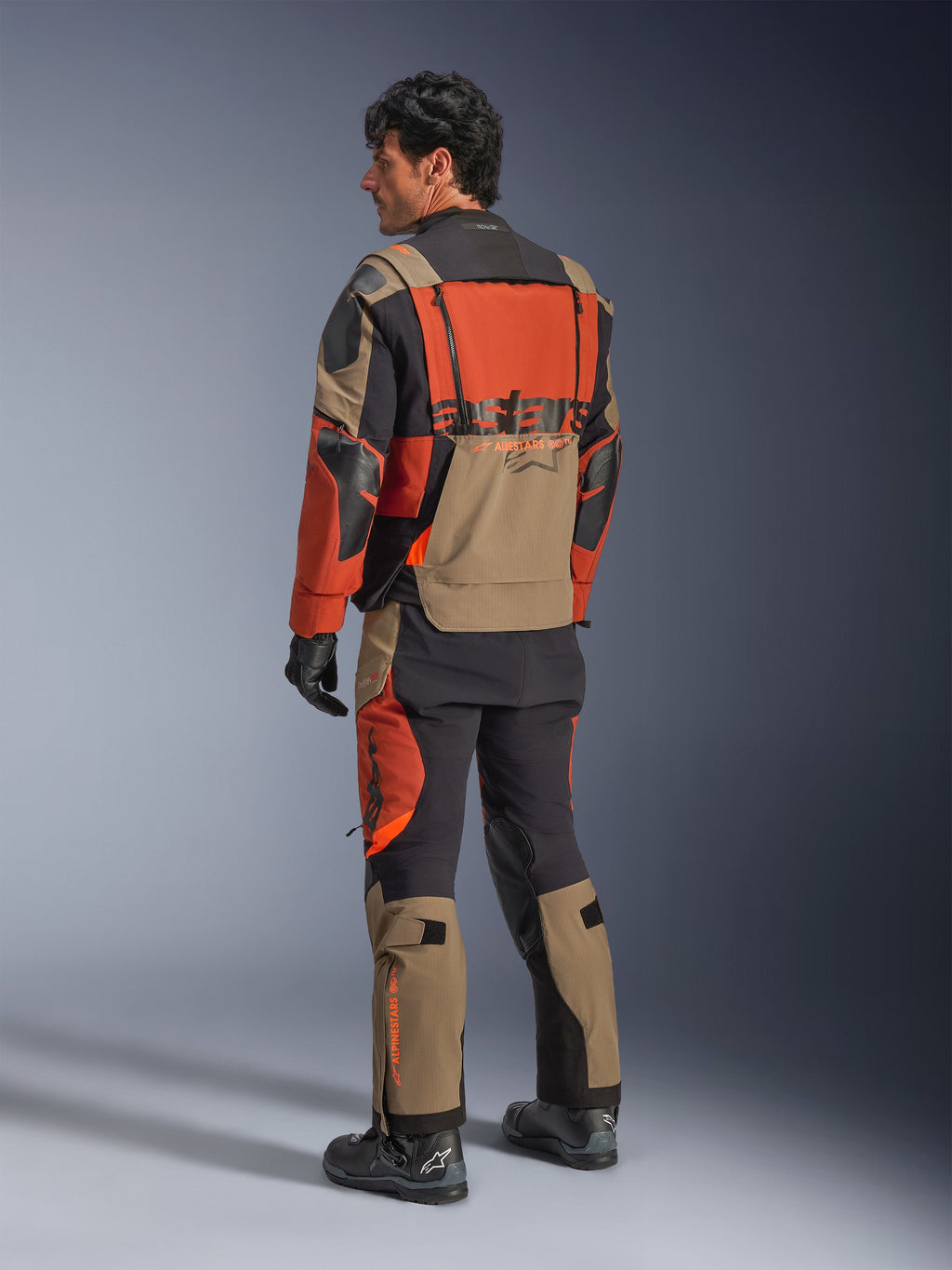 Giacca laminata Alpinestars Halo Pro Drystar® XF, giacca da moto adventure Walnut Rust Black, vista posteriore indossata con pantaloni coordinati e stivali neri, caratterizzata da materiale tecnico stretch 3L, pannelli di ventilazione modulari e branding arancione sul retro.