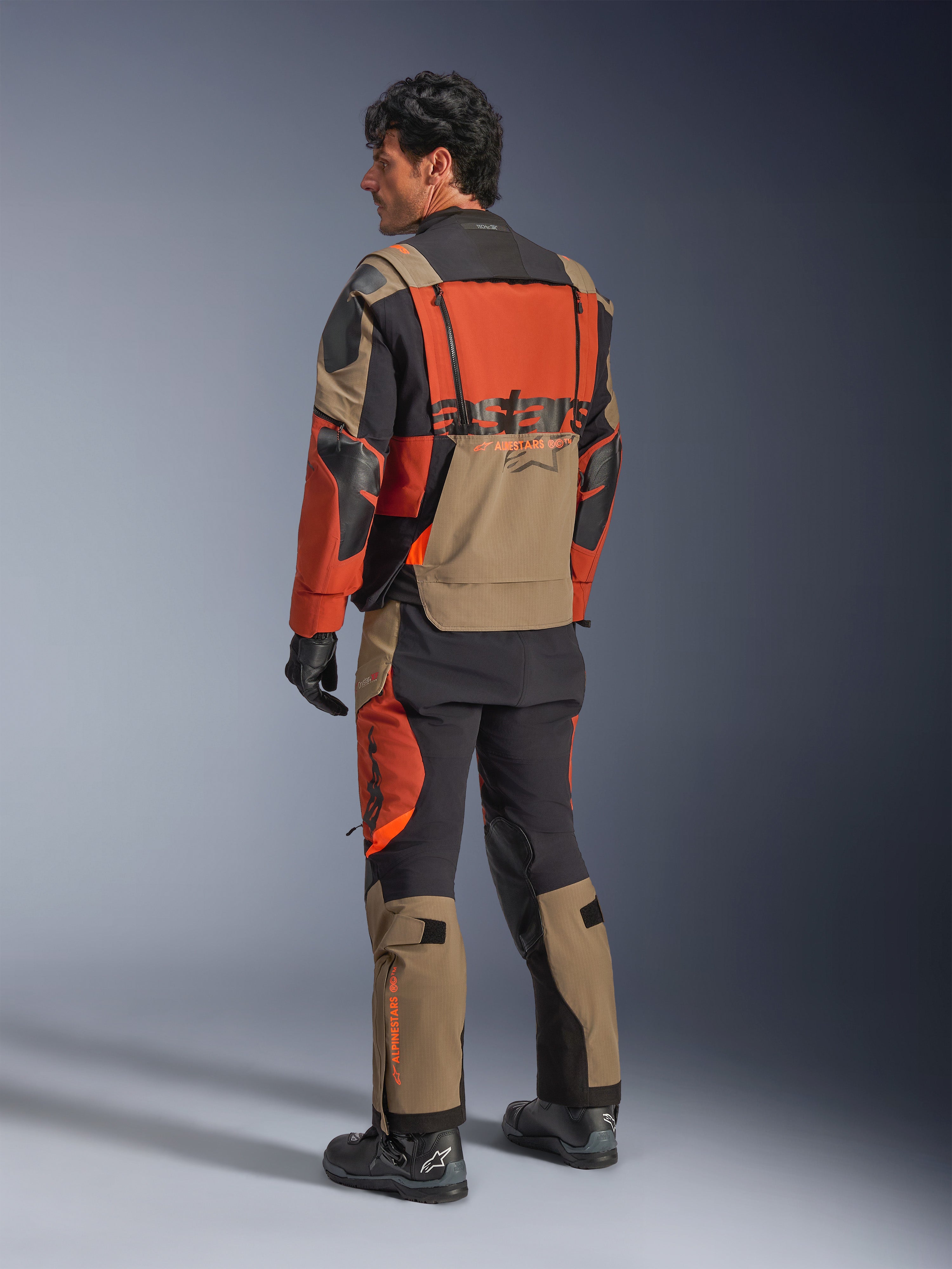 Giacca laminata Alpinestars Halo Pro Drystar® XF, giacca da moto adventure Walnut Rust Black, vista posteriore indossata con pantaloni coordinati e stivali neri, caratterizzata da materiale tecnico stretch 3L, pannelli di ventilazione modulari e branding arancione sul retro.