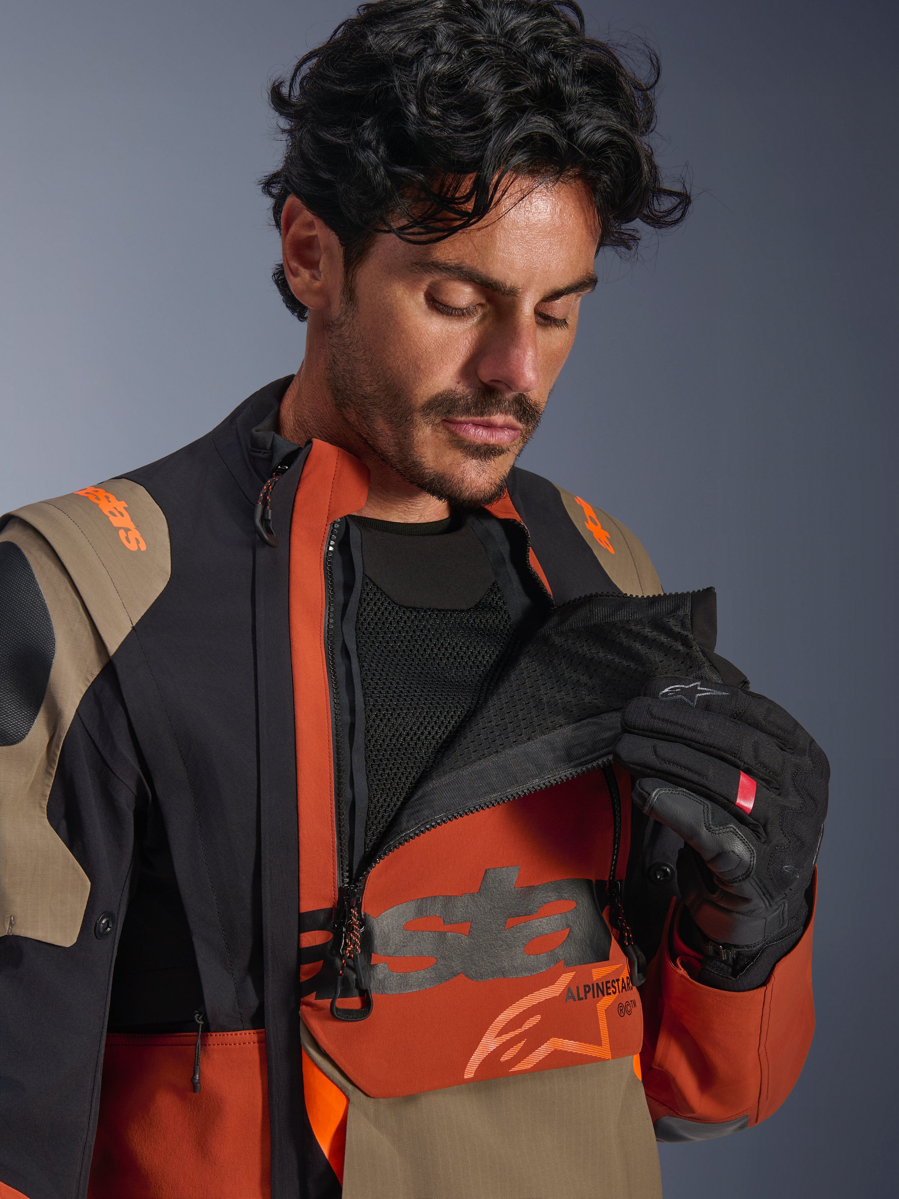 Giacca laminata Alpinestars Halo Pro Drystar® XF, colorazione Walnut Rust Black, modello maschile che dimostra il sistema di ventilazione modulare aprendo il pannello frontale sul petto per rivelare la rete nera, giacca tecnica da moto adventure con accenti ruggine e sabbia.