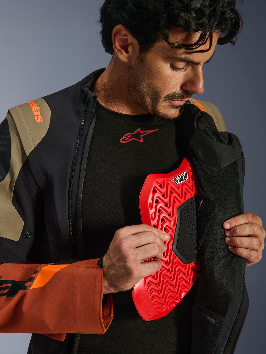 Alpinestars Halo Pro Drystar® XF Laminated Jacket in Walnut Rust Black, scatto lifestyle di un modello che apre la zip della giacca adventure per mostrare un inserto protettivo per il torace rosso, con marchio arancione sulla spalla e base layer nero con logo rosso.