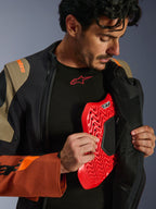 Alpinestars Halo Pro Drystar® XF Laminated Jacket in Walnut Rust Black, scatto lifestyle di un modello che apre la zip della giacca adventure per mostrare un inserto protettivo per il torace rosso, con marchio arancione sulla spalla e base layer nero con logo rosso.