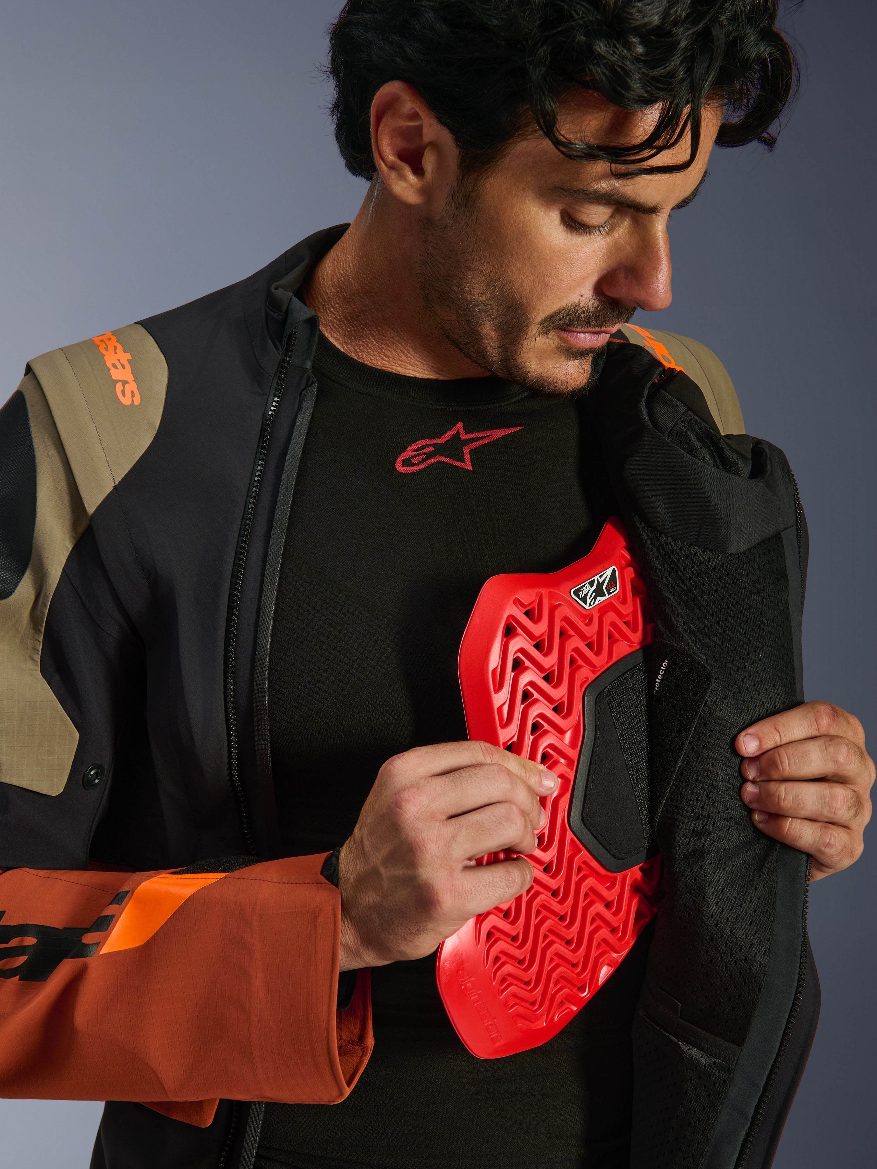 Alpinestars Halo Pro Drystar® XF Laminated Jacket in Walnut Rust Black, scatto lifestyle di un modello che apre la zip della giacca adventure per mostrare un inserto protettivo per il torace rosso, con marchio arancione sulla spalla e base layer nero con logo rosso.