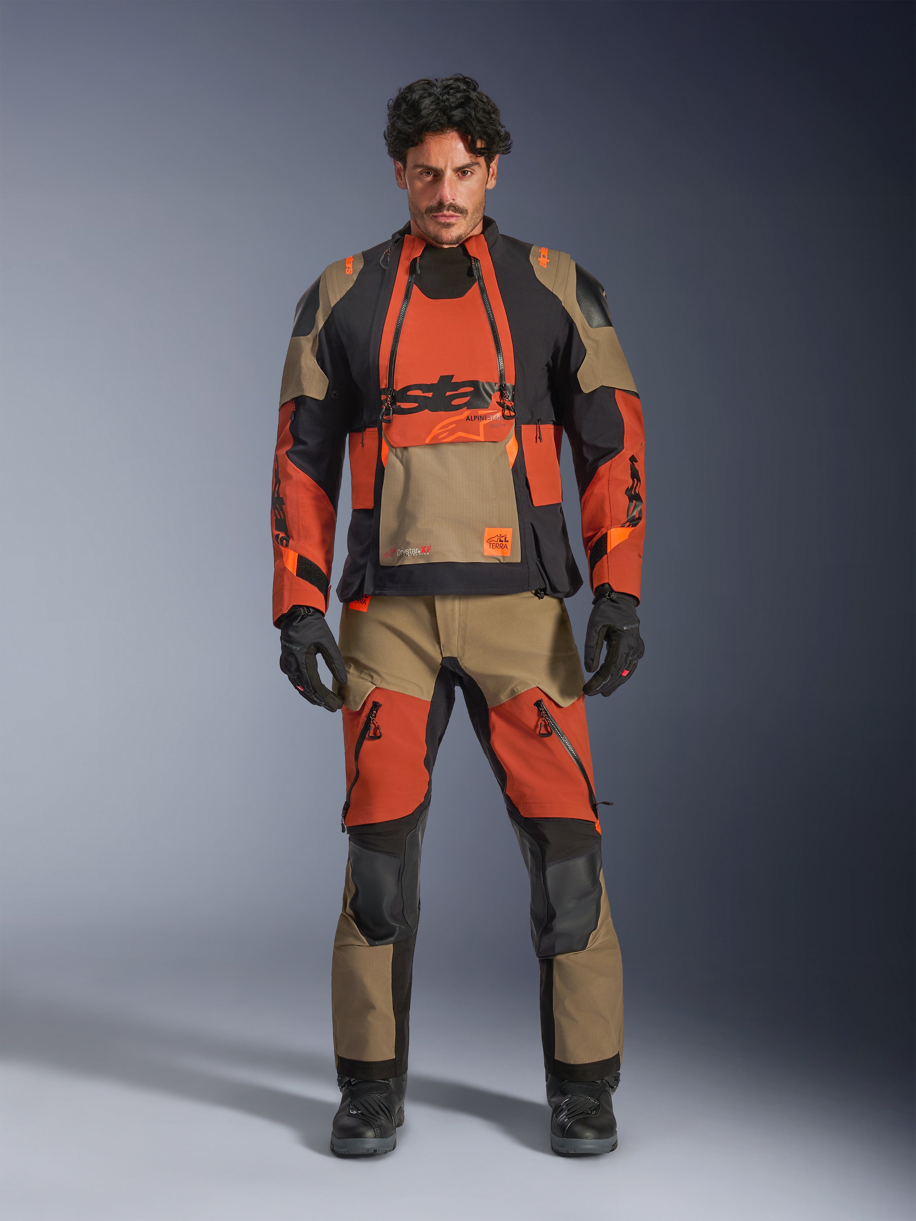 Modello che indossa la giacca laminata Alpinestars Halo Pro Drystar® XF e pantaloni coordinati nella colorazione Walnut Rust Black, abbigliamento modulare da moto adventure con pannelli di ventilazione frontali e cerniere, completato da guanti e stivali da moto neri.