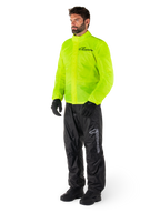 Uomo che indossa la giacca Alpinestars Hurricane Rain V2 in Giallo Fluo e i pantaloni Hurricane Rain V2 neri, abbigliamento antipioggia da moto impermeabile e ad alta visibilità, con loghi riflettenti su petto e coscia, outfit completo con guanti e stivali neri.