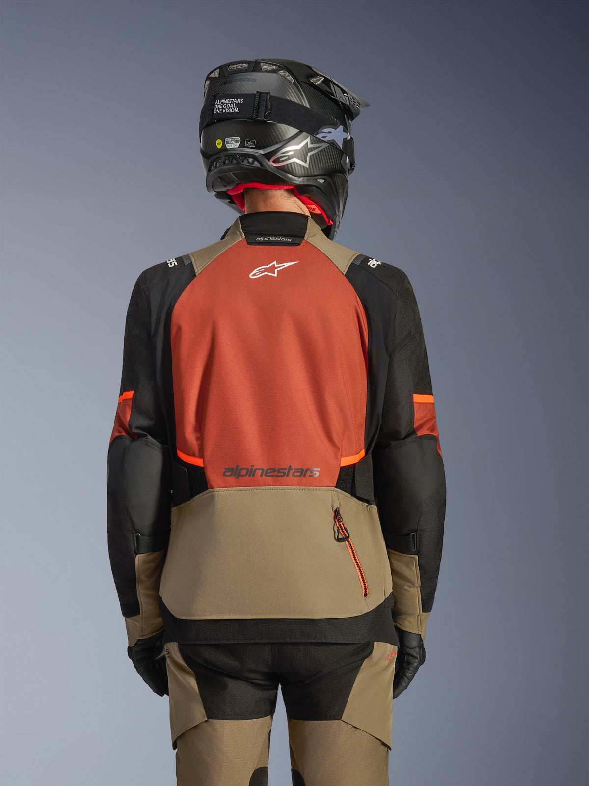 Vista posteriore della giacca Alpinestars Andes V4 Drystar® in Walnut Rust Black, giacca da touring caratterizzata da un ampio pannello posteriore color ruggine con logo bianco, sezione inferiore noce con tasca a cerniera verticale e maniche nere con accenti arancioni, abbinata a pantaloni coordinati e casco nero per la guida in ogni condizione atmosferica.