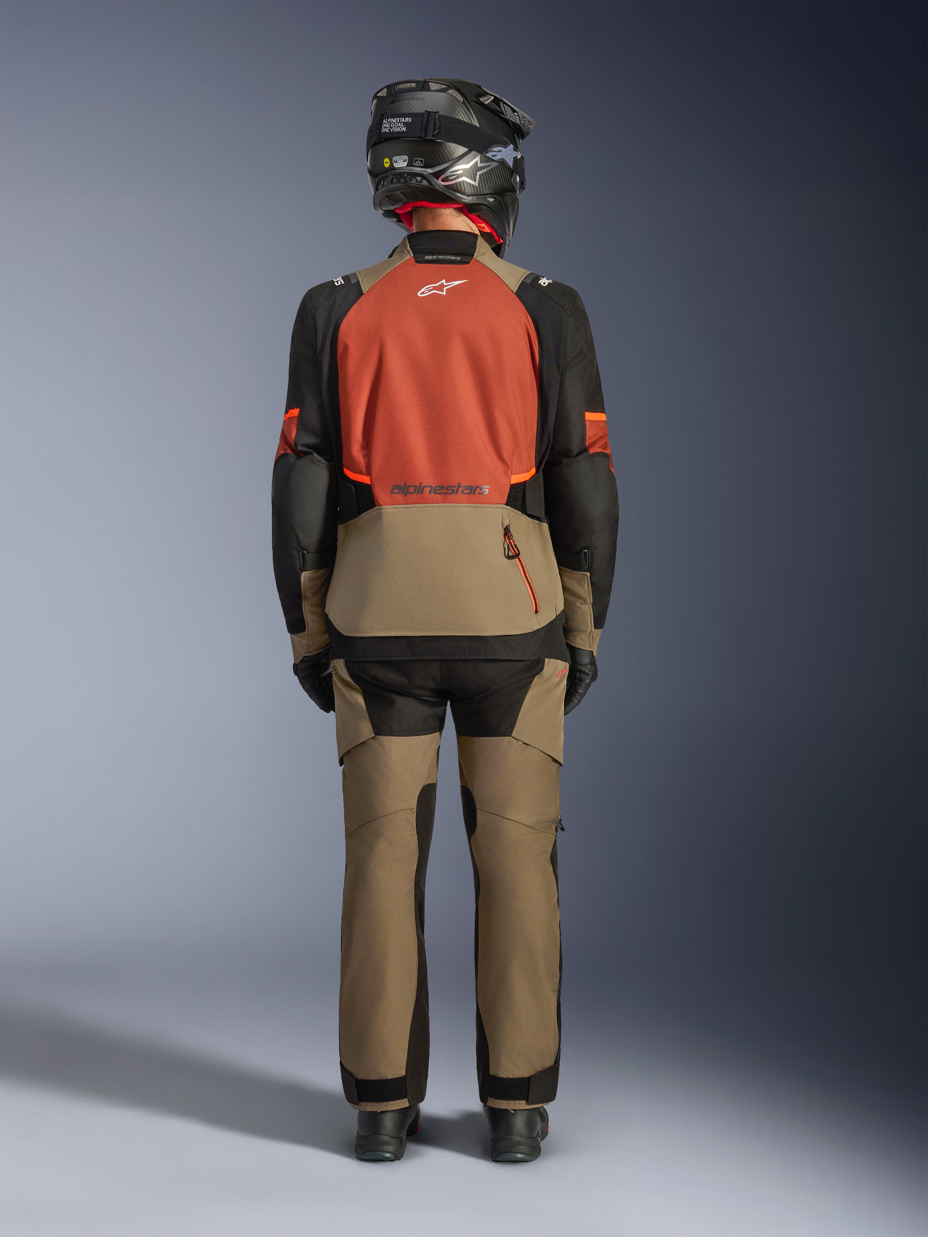 Vista posteriore di un motociclista che indossa la giacca Alpinestars Andes V4 Drystar® in Walnut Rust Black, abbinata a pantaloni adventure coordinati e un casco nero, mettendo in risalto il pannello posteriore color ruggine, l'ampia tasca posteriore e i loghi riflettenti.