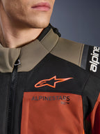 Giacca Alpinestars Andes V4 Drystar® in Walnut Rust Black, primo piano della parte superiore del petto e della spalla, con logo A-star arancione su pannello nero e marchio Alpinestars sulla sezione color ruggine, dettaglio dell'equipaggiamento da moto adventure