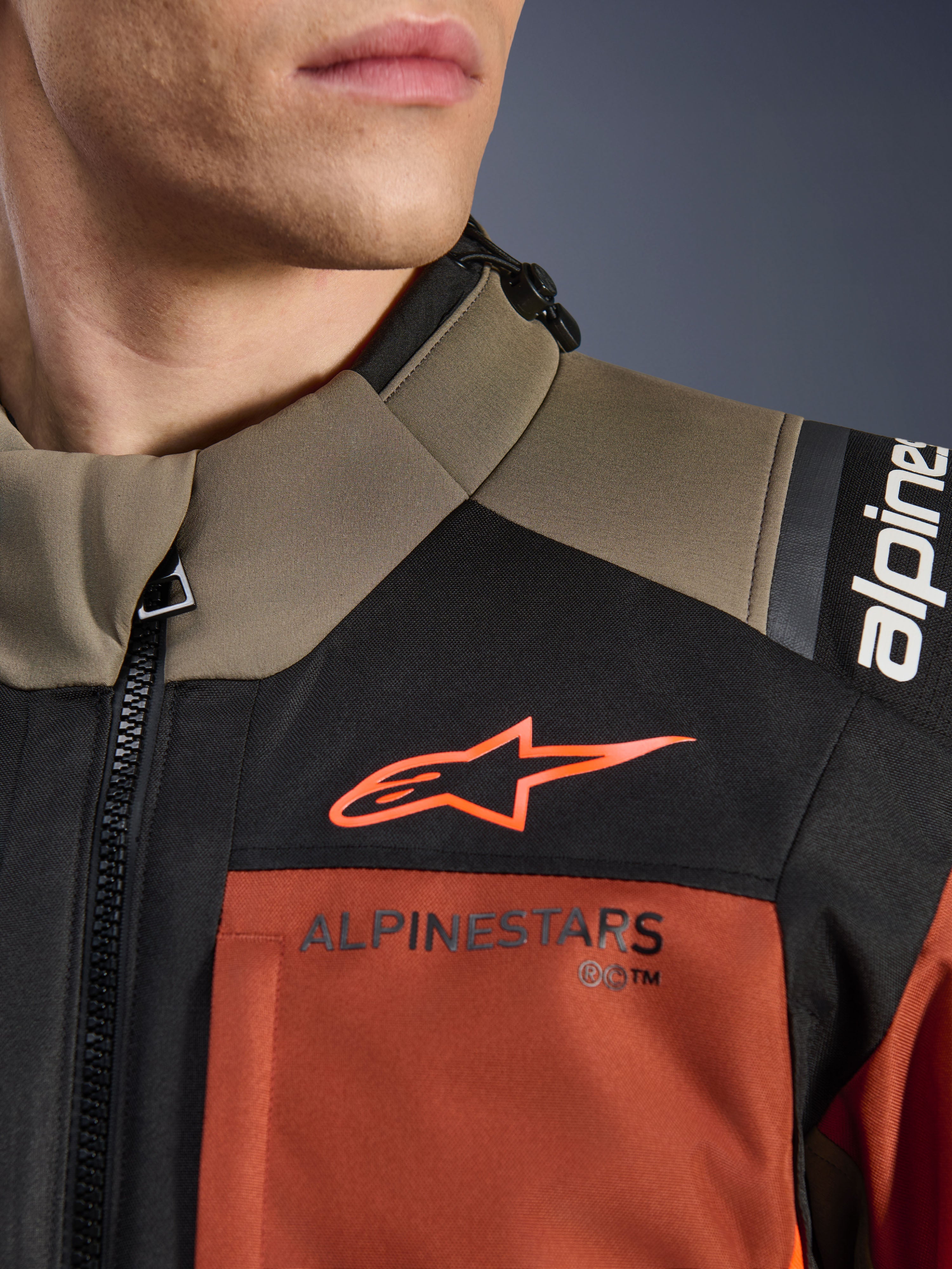 Giacca Alpinestars Andes V4 Drystar® in Walnut Rust Black, primo piano della parte superiore del petto e della spalla, con logo A-star arancione su pannello nero e marchio Alpinestars sulla sezione color ruggine, dettaglio dell'equipaggiamento da moto adventure