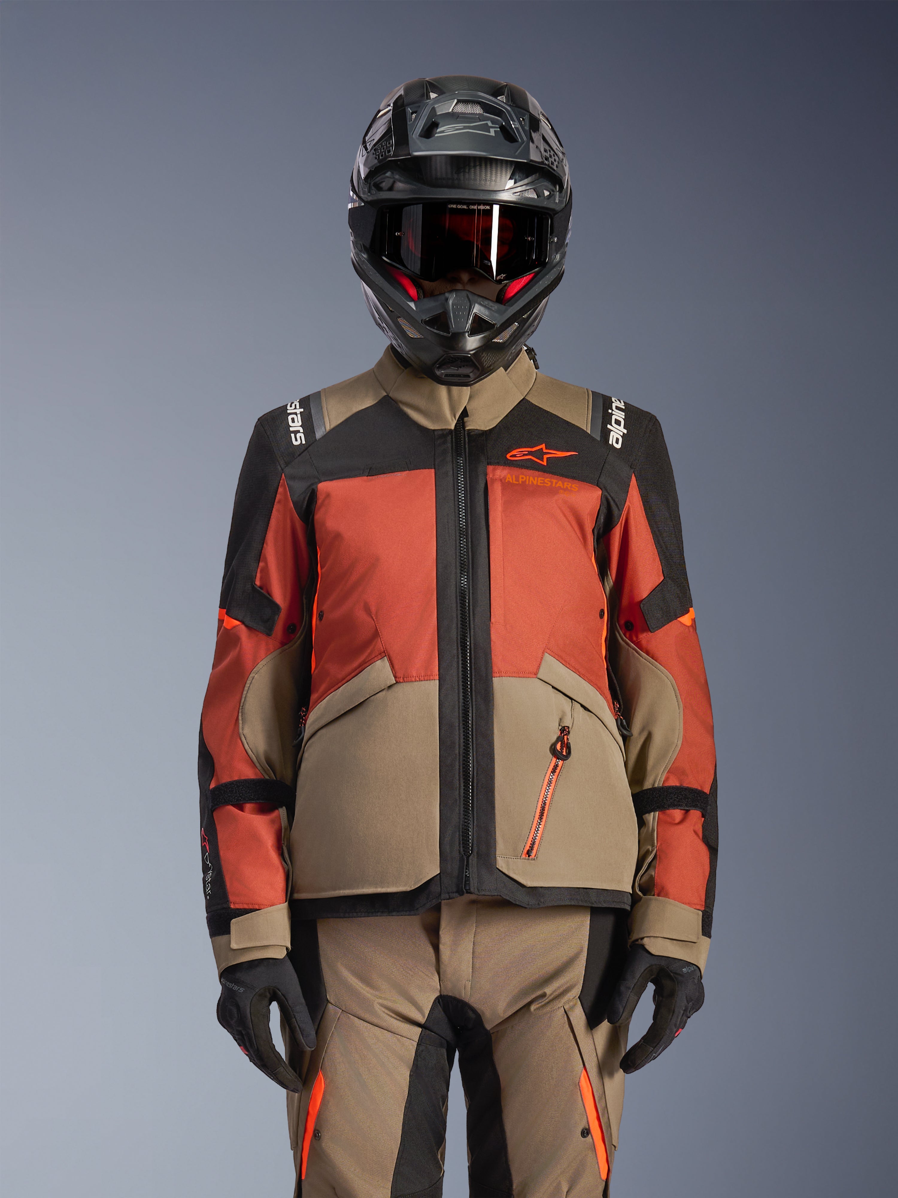 Giacca Alpinestars Andes V4 Drystar®, giacca da moto adventure, Walnut Rust Black, indossata con casco e guanti coordinati, caratterizzata da un design tecnico a pannelli multipli in sabbia, arancione-rossastro e nero con logo arancione visibile e cerniere impermeabili.