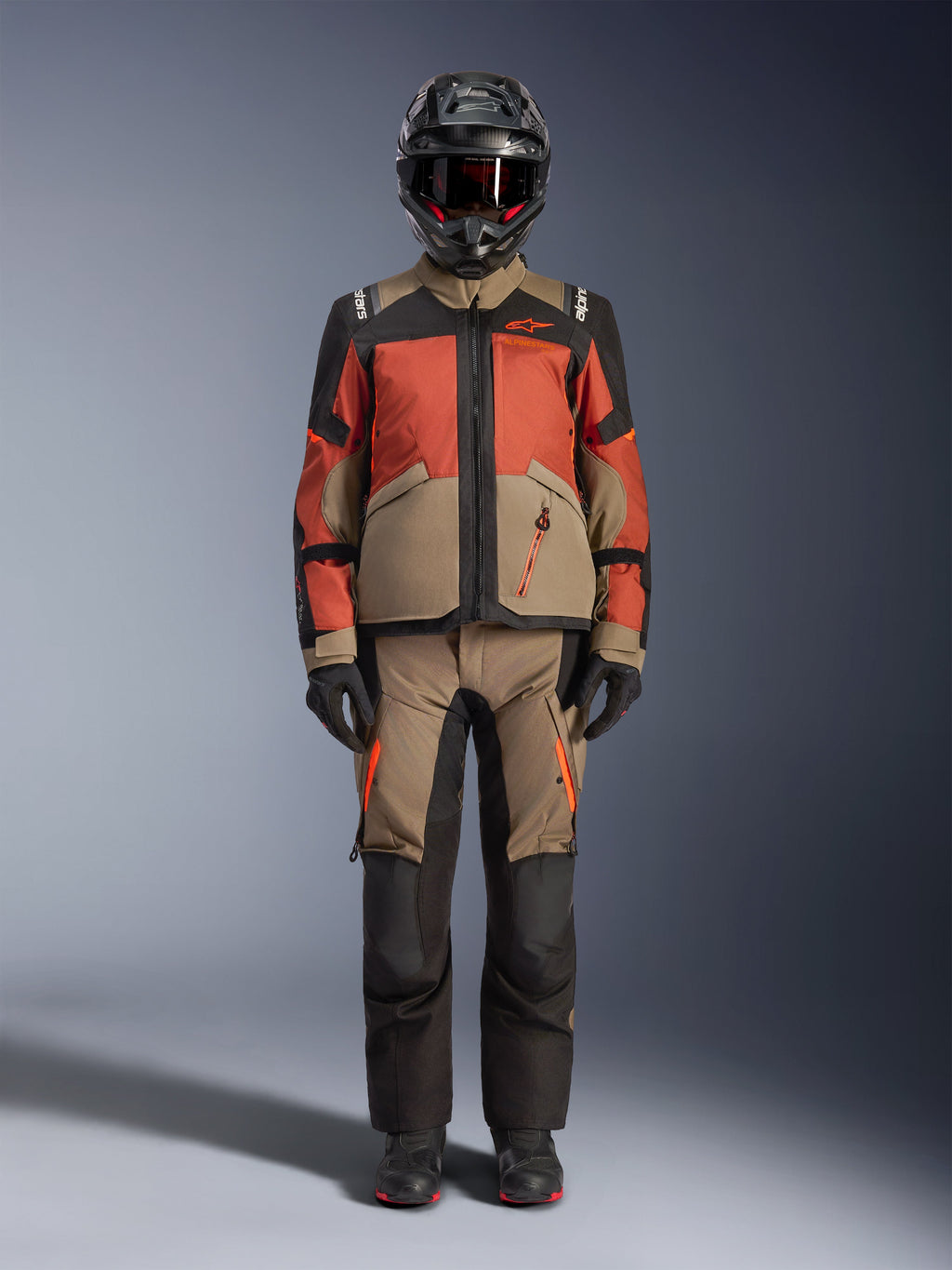 Pilota adventure che indossa la giacca Alpinestars Andes V4 Drystar® in Walnut Rust Black, abbinata a pantaloni adventure coordinati, casco nero con dettagli rossi e maschera, e guanti neri, che mostra un outfit completo da touring per ogni condizione atmosferica.