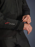 Giacca Alpinestars Andes V4 Drystar®, giacca da moto in tessuto nero, primo piano della manica con logo Alpinestars rosso e branding Drystar® bianco, pilota che regola il cinturino in velcro sul polso per una vestibilità sicura, dettaglio funzionale per la guida adventure