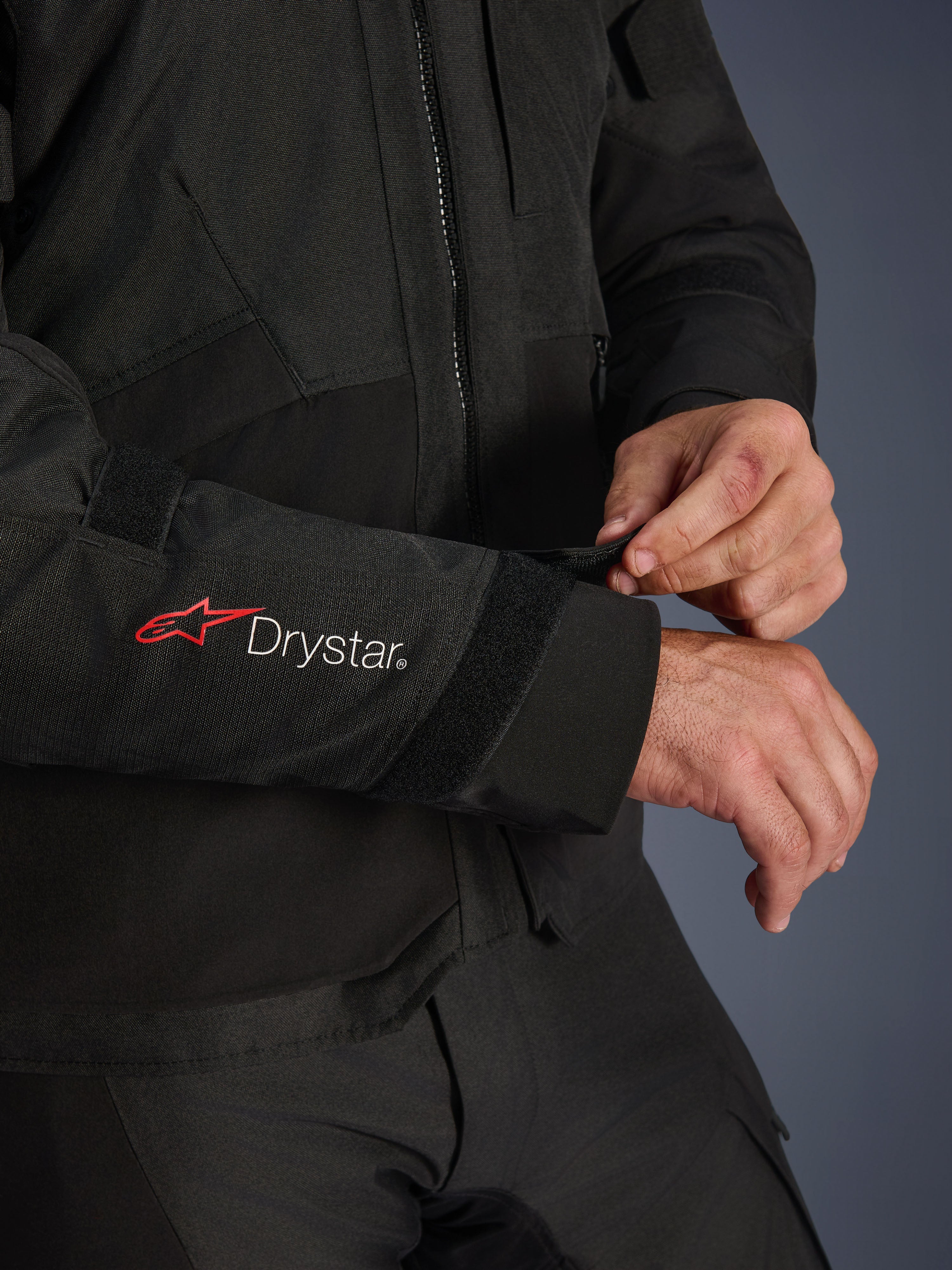 Giacca Alpinestars Andes V4 Drystar®, giacca da moto in tessuto nero, primo piano della manica con logo Alpinestars rosso e branding Drystar® bianco, pilota che regola il cinturino in velcro sul polso per una vestibilità sicura, dettaglio funzionale per la guida adventure