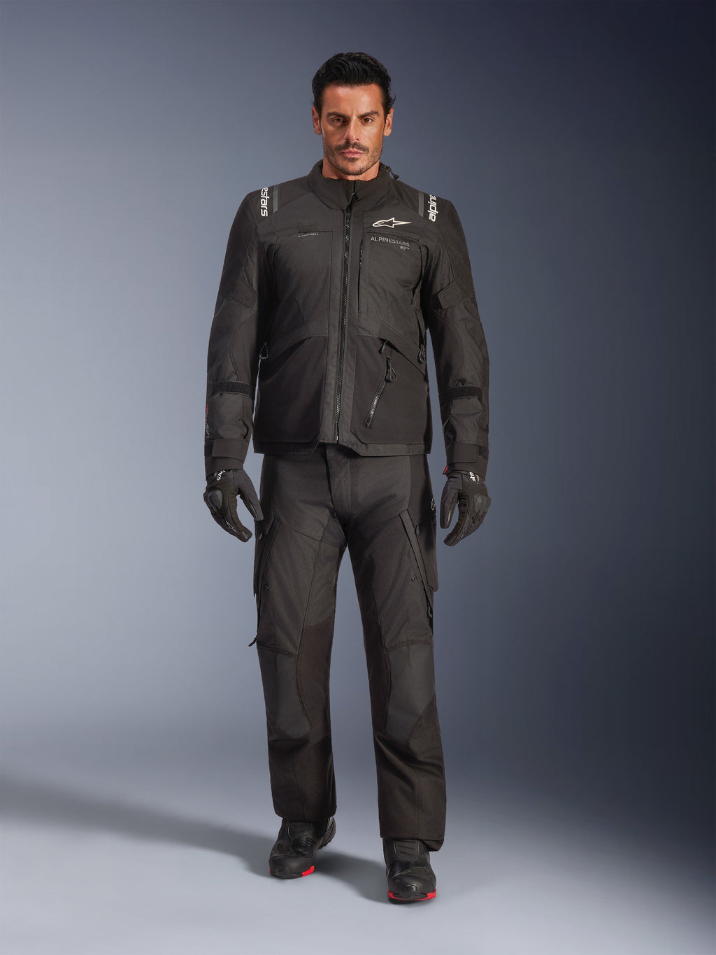 Modello che indossa la giacca Alpinestars Andes V4 Drystar® in nero, abbinata a pantaloni da moto adventure coordinati, guanti neri e stivali neri, scatto in studio a figura intera che mostra l'equipaggiamento tecnico da touring per tutte le stagioni con marchio Alpinestars.