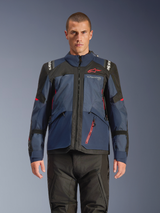 Alpinestars Giacca Andes V4 Drystar®, giacca da moto adventure, colori Night Navy e Mood Indigo con pannelli neri, indossata da un modello con pantaloni tecnici neri, logo Alpinestars rosso sul petto e dettaglio zip rossa, struttura in tessuto resistente per il touring in ogni condizione climatica.
