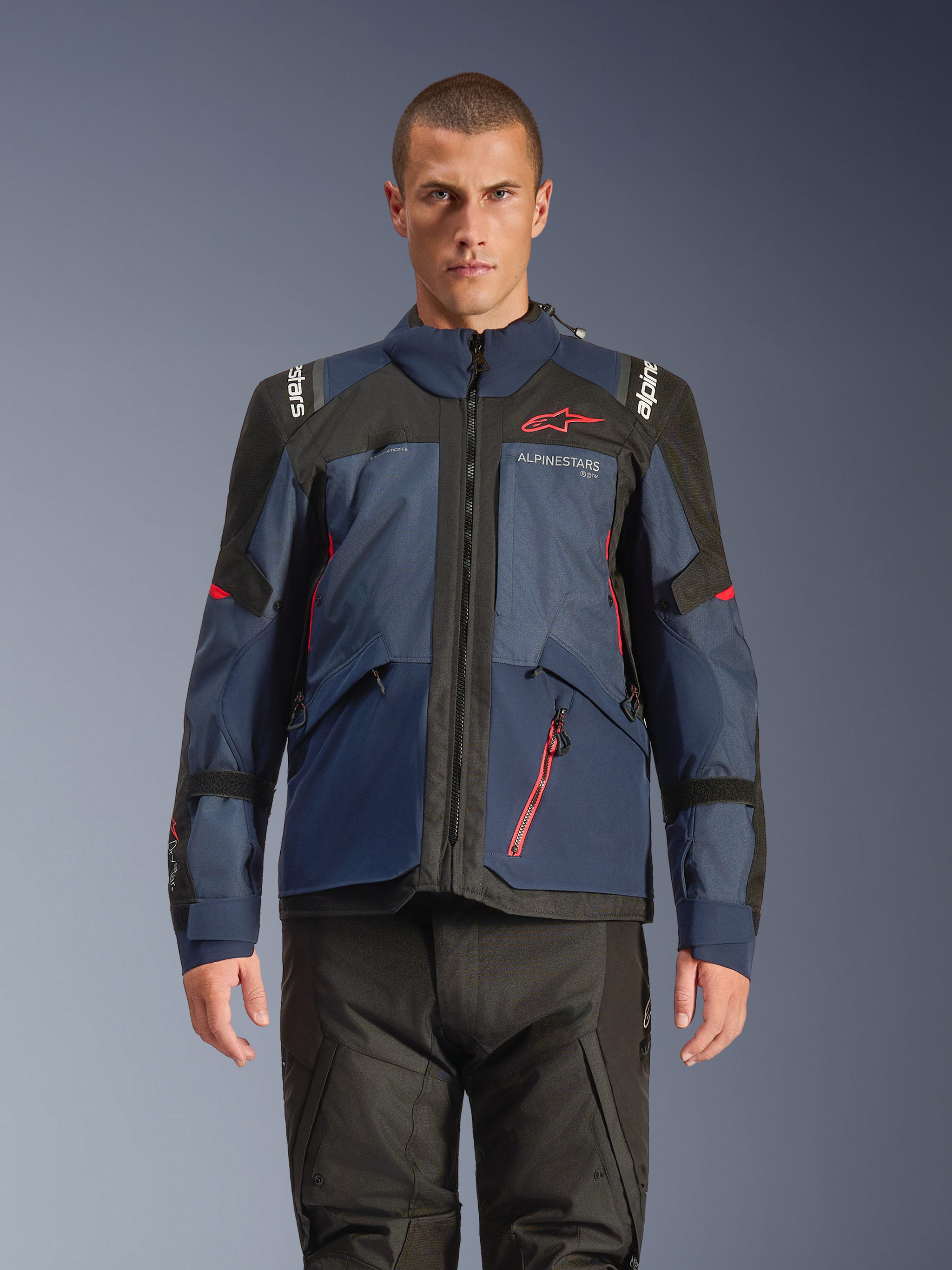 Alpinestars Giacca Andes V4 Drystar®, giacca da moto adventure, colori Night Navy e Mood Indigo con pannelli neri, indossata da un modello con pantaloni tecnici neri, logo Alpinestars rosso sul petto e dettaglio zip rossa, struttura in tessuto resistente per il touring in ogni condizione climatica.
