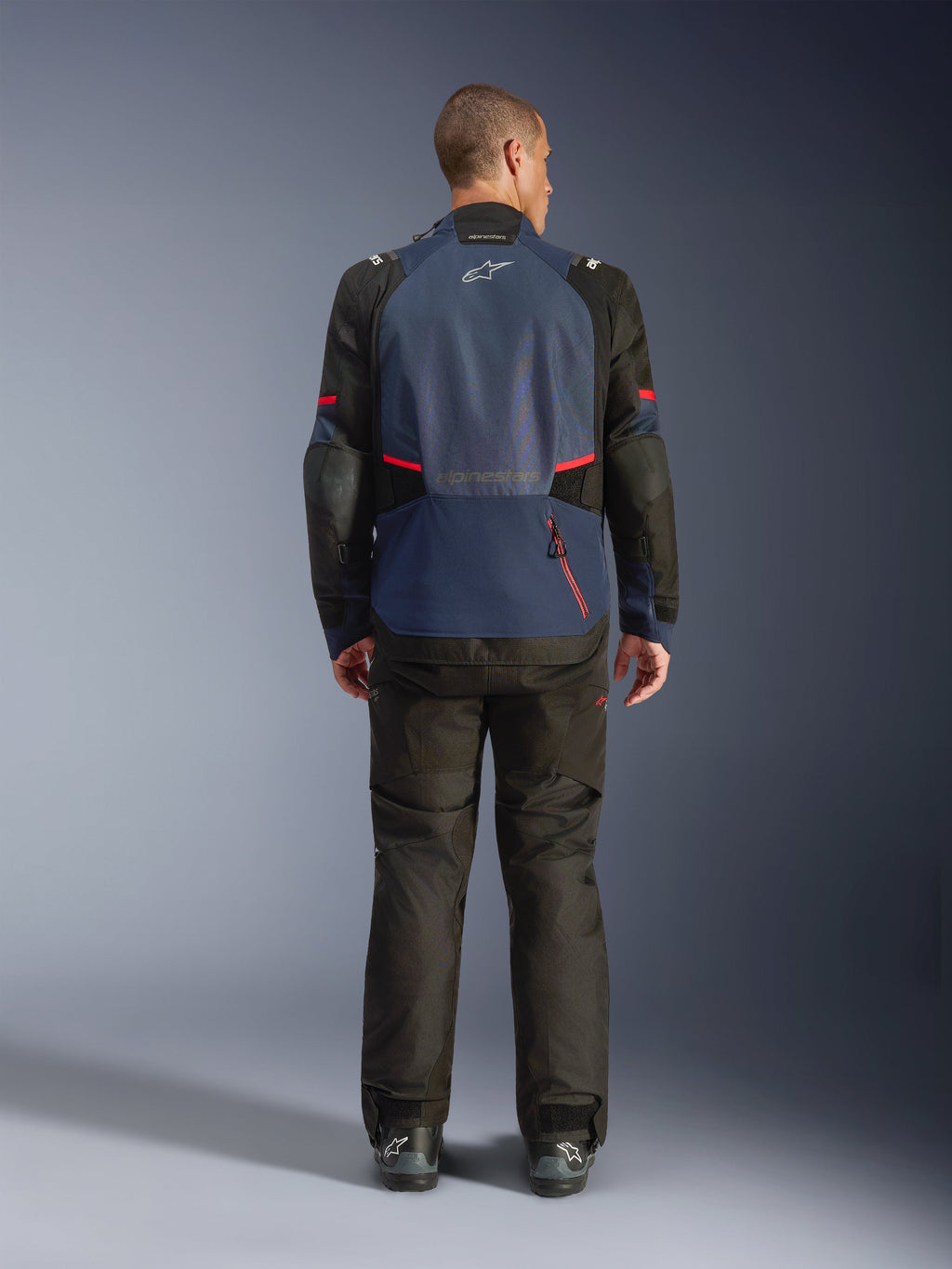 Giacca Alpinestars Andes V4 Drystar®, vista posteriore, colori Night Navy e Mood Indigo con pannelli neri e dettagli rossi, indossata con pantaloni neri abbinati, con logo Alpinestars sulla parte superiore della schiena e cerniera per l'ampio scomparto posteriore.